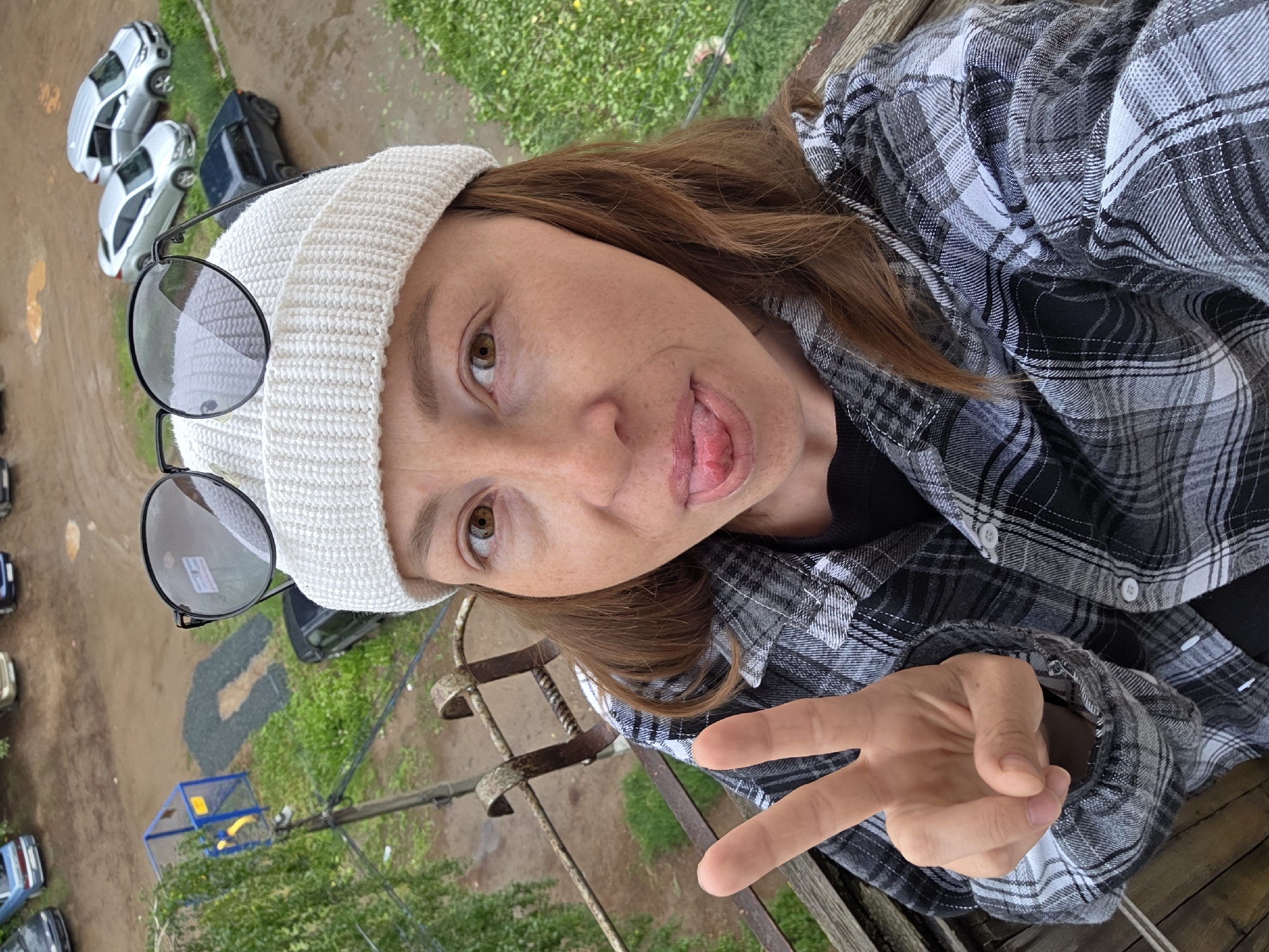 A woman takes a selfie from an elevated position, looking down over an outdoor scene in Chaikovsky, Russia. She is depicted from the chest up, positioned diagonally across the frame with her head tilted. She has reddish-brown hair and light brown eyes, and is wearing a white knit beanie, with round, dark-rimmed glasses perched on top of it. Her attire includes a black and white plaid button-up shirt over a dark undershirt. Her facial expression is playful, with her tongue slightly sticking out. Her left hand is raised, showing a peace sign.
The background, viewed from above, features a ground covered in patches of green grass and damp, brownish-grey dirt or mud, suggesting recent rain. Several parked cars are visible below, including at least two silver sedans and a dark SUV. In the middle ground, there is a distinct dark, 'G' or 'C' shaped marking on the ground. A blue and yellow structure, possibly a small playground element or utility box, is also seen, along with a utility pole and overhead wires. A rusty metal railing, likely part of a balcony or window, is prominent in the foreground, indicating the height from which the photo was taken. The overall lighting suggests it is daytime, possibly overcast. No discernible text is visible in the image.