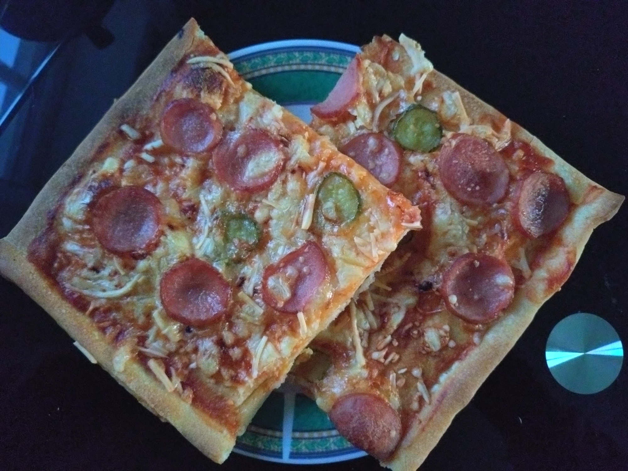Two slices of pepperoni pizza are on a dark, reflective surface. The pizza slices are square-cut and overlap each other. Both slices are topped with tomato sauce, cheese, pepperoni slices, and pieces of pickle. The crust of the pizza is golden brown and appears to be thin. Underneath the pizza slices is a plate with a green and blue patterned rim. To the right of the pizza, there is a circular, metallic object that reflects a blue light. The background is dark and out of focus, suggesting the image was taken indoors, possibly in a kitchen or dining area. There are no people visible in the image. There is no information in the image to suggest the location in the city of Averøy, Norway.