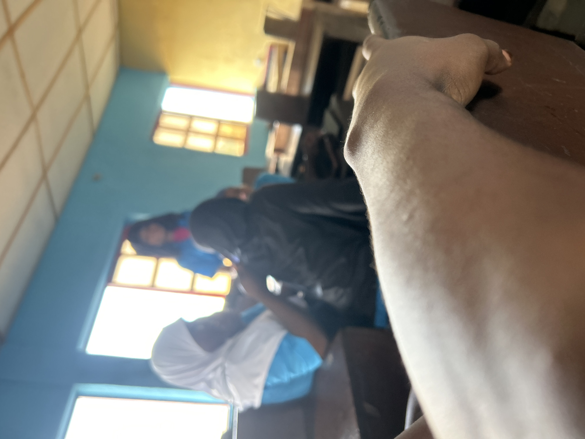 The image depicts an indoor scene in Ikorodu, Nigeria, shot from a low, rotated perspective, showing a classroom or similar learning environment during the daytime. In the immediate foreground, a person's bare forearm and hand with dark skin rest on a dark brown wooden desk or table, dominating the right side of the frame.
The background, largely out of focus, reveals several individuals. To the left, a person wearing a white head covering (possibly a hijab) and a light blue top is visible, seemingly looking downwards. Behind them, another individual wears a dark hoodie or jacket. Further back and to the right, a person with dark hair is seen wearing a blue top, near a brightly lit window.
The room features light blue walls and a ceiling with a rectangular grid pattern. Several large windows are present, letting in abundant natural light, indicating it is daytime. The bright light from the windows creates strong highlights and some lens flare. Behind the windows, a warmer, yellow-toned wall or light source is discernible. There are also hints of other furniture or shelves in the blurry background.
The overall scene suggests a quiet gathering, possibly a class or study session, with participants seated at desks. No specific activity or emotion is clearly discernible due to the depth of field and rotation, but the setting implies a learning or assembly context. There is no visible text in the image.