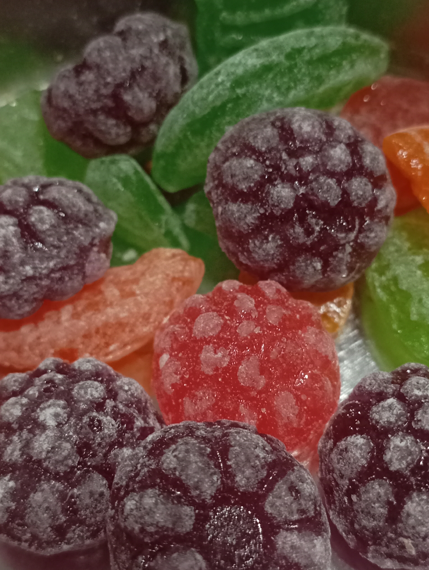 The image shows a close-up of various hard candies. The candies are of different shapes and colors, including round, bumpy shapes resembling raspberries or blackberries in dark purple, and elongated shapes in green and orange. A red round candy is also present. The candies appear to have a slightly frosted or sugary coating. There is no indication of the location being Krasnodar, Russia, or any people, subjects, objects, actions, interactions, or environment beyond the candies themselves.