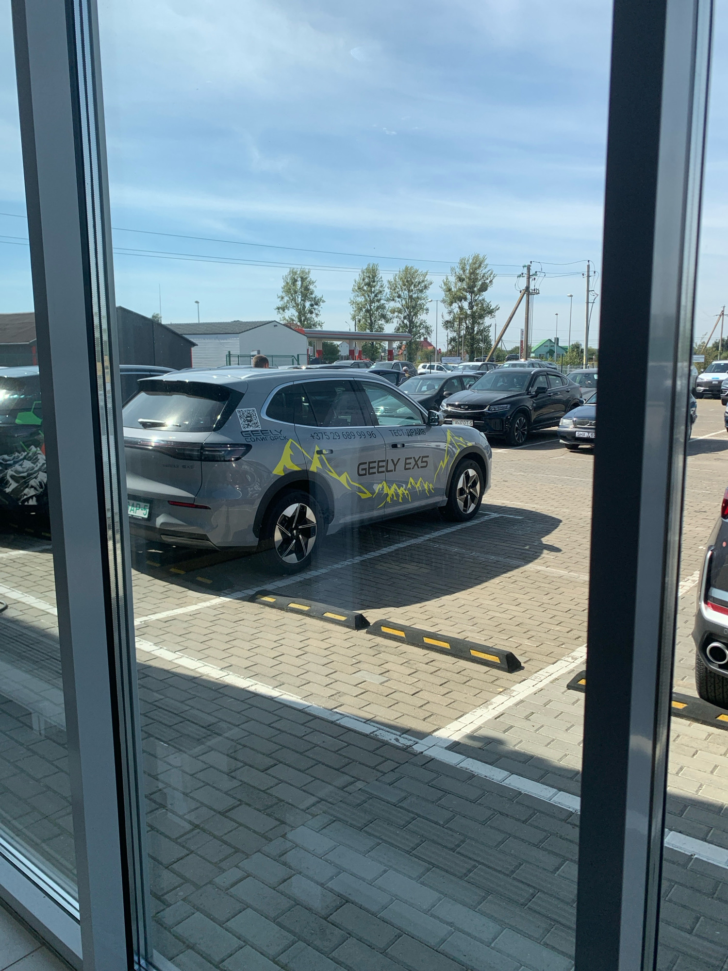 The image shows a gray Geely EX5 SUV parked in a lot, viewed through a glass storefront. The SUV has yellow graphic accents and text on its side, including "GEELY EX5" and a phone number, "+375 29 689 99 96". A QR code is also visible on the rear of the vehicle, along with text in Cyrillic that appears to be a city name. The parking lot is paved with light-colored bricks and has yellow and black parking wheel stops. Other cars are visible in the background, including a black sedan and a portion of a white SUV. In the distance, there are buildings, trees, and power lines, suggesting an outdoor commercial or industrial area. The sky is blue with scattered clouds, indicating daylight. The camera is positioned at an angle looking out from inside a building with a dark gray frame in the foreground. There are no people clearly identifiable in the image, though a person's head is partially visible over the roof of the black sedan in the background. The context suggests this is likely a car dealership or an area with a concentration of vehicles. Based on the Cyrillic text and phone number format, it is plausible this scene is located in Belarus, possibly in or near the city of Чыжэвіцкі сельскі Савет.