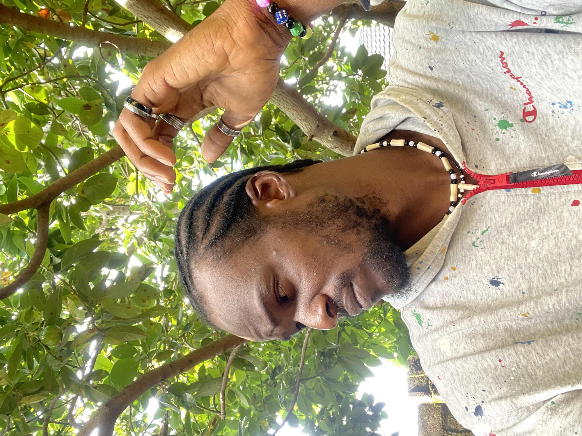 A Black man with dark skin, braids, and a goatee is pictured in a medium shot, looking to his left. He wears a grey Champion hoodie with colorful paint splatters, a beaded necklace, and several rings on his right hand, including a silver band and a patterned ring. A colorful beaded bracelet with dice charms adorns his right wrist. He is outdoors, partially obscured by the branches and green leaves of a tree, possibly an avocado tree, with a bright sky visible in the background. In the lower left corner, a brick wall is visible. The scene is likely located in Jos, Nigeria, suggested by the characteristic foliage and architectural elements. The man's hand is raised to his head, with his fingers gently touching his hair or earrings.