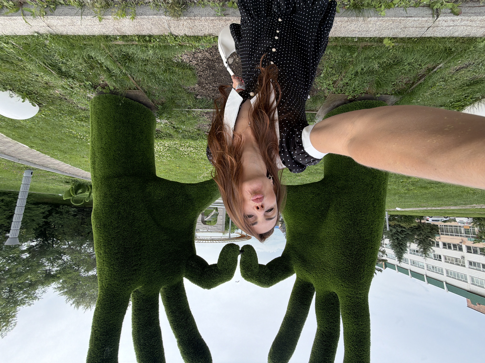 In Birobidzhan, Russia, a woman is taking a selfie in front of two large, green, topiary sculptures shaped like hands forming a heart. The woman has long, brown hair and is wearing a black and white polka dot dress with a white collar. She is puckering her lips for the photo and holding a white purse. The sculptures are located in a park-like setting with grass, trees, and a paved walkway. In the background, there is a large white building with green trim. The sky is overcast.