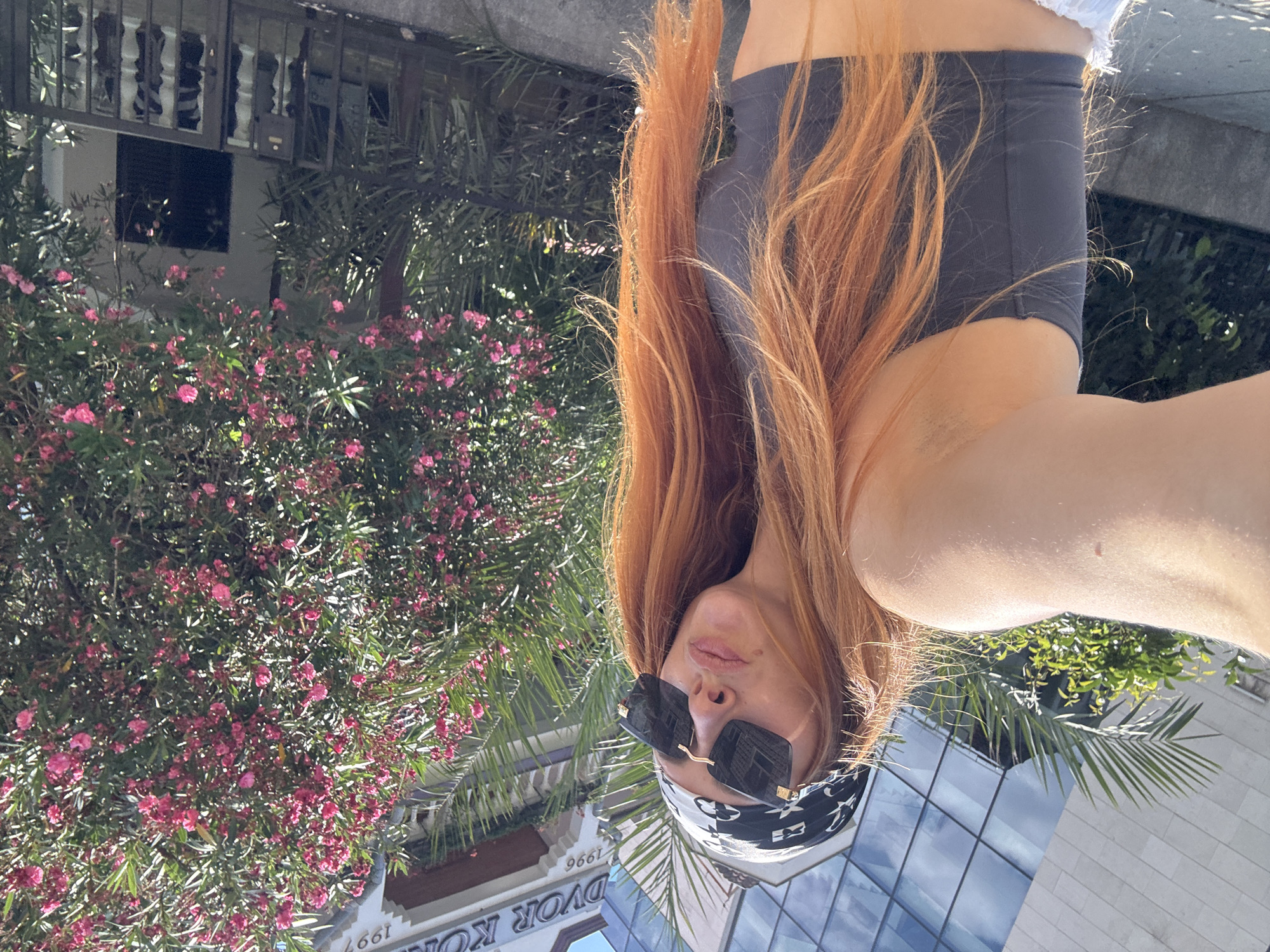 The image shows a young woman with long, reddish-blonde hair, wearing sunglasses and a bandana. She is outdoors, with a building and lush greenery in the background. The setting appears to be a sunny day, likely in the morning or afternoon, given the bright lighting.
In the foreground, the woman is partially visible, with her face turned towards the camera and her hair falling around her. She's wearing dark-colored shorts and a white top. Her sunglasses have dark lenses and a square frame, and the bandana is black and white with a repeating pattern.
The background features a vibrant bush with numerous small pink flowers, likely oleander. Behind the flowers, a building with architectural details, including latticed windows, is visible. Further back, a modern building with a glass facade reflects the sky. Palm fronds from a nearby tree are also visible.
The overall impression is of a relaxed, summery scene, possibly in a tourist area given the architecture and landscaping. There is some visible text on the building in the lower part of the image, which appears to read "DVOR KON" and "1996". The image is oriented sideways.