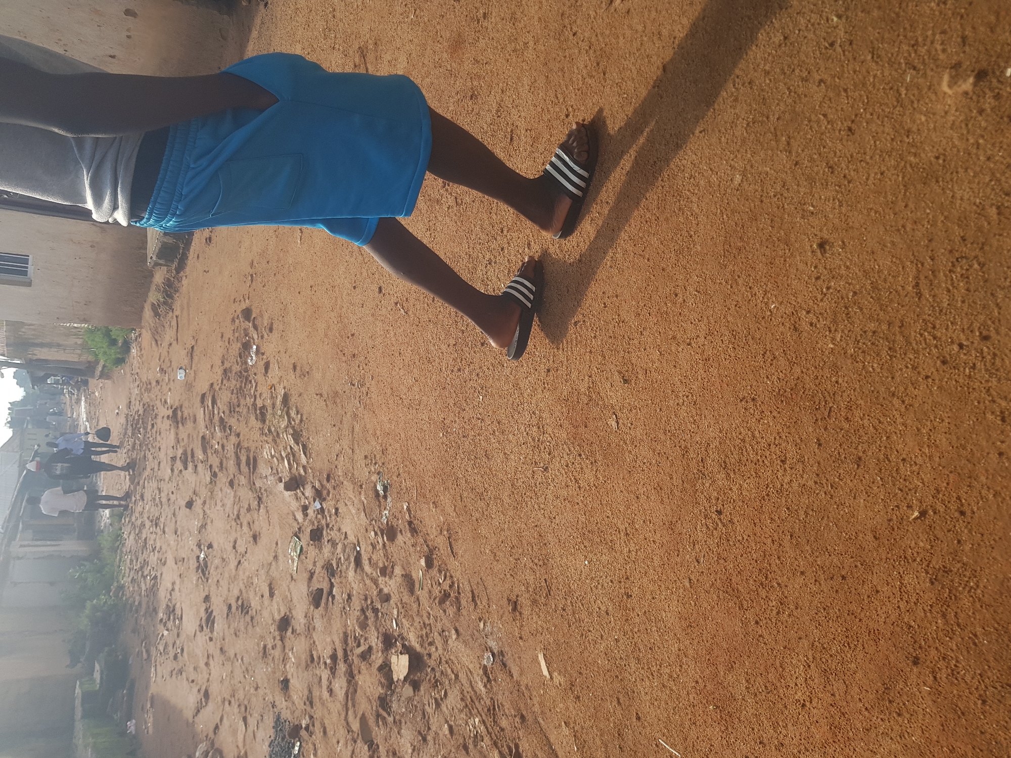 A person is standing with their hands in their pockets and wearing blue shorts and black and white sandals on a dirt road in Jos, Nigeria. The road is unpaved and covered with reddish-brown dirt and scattered debris. In the background, there are buildings with corrugated metal roofs and what appear to be homes. Two other people are visible walking further down the road. The scene suggests a typical street in a residential area of Jos.