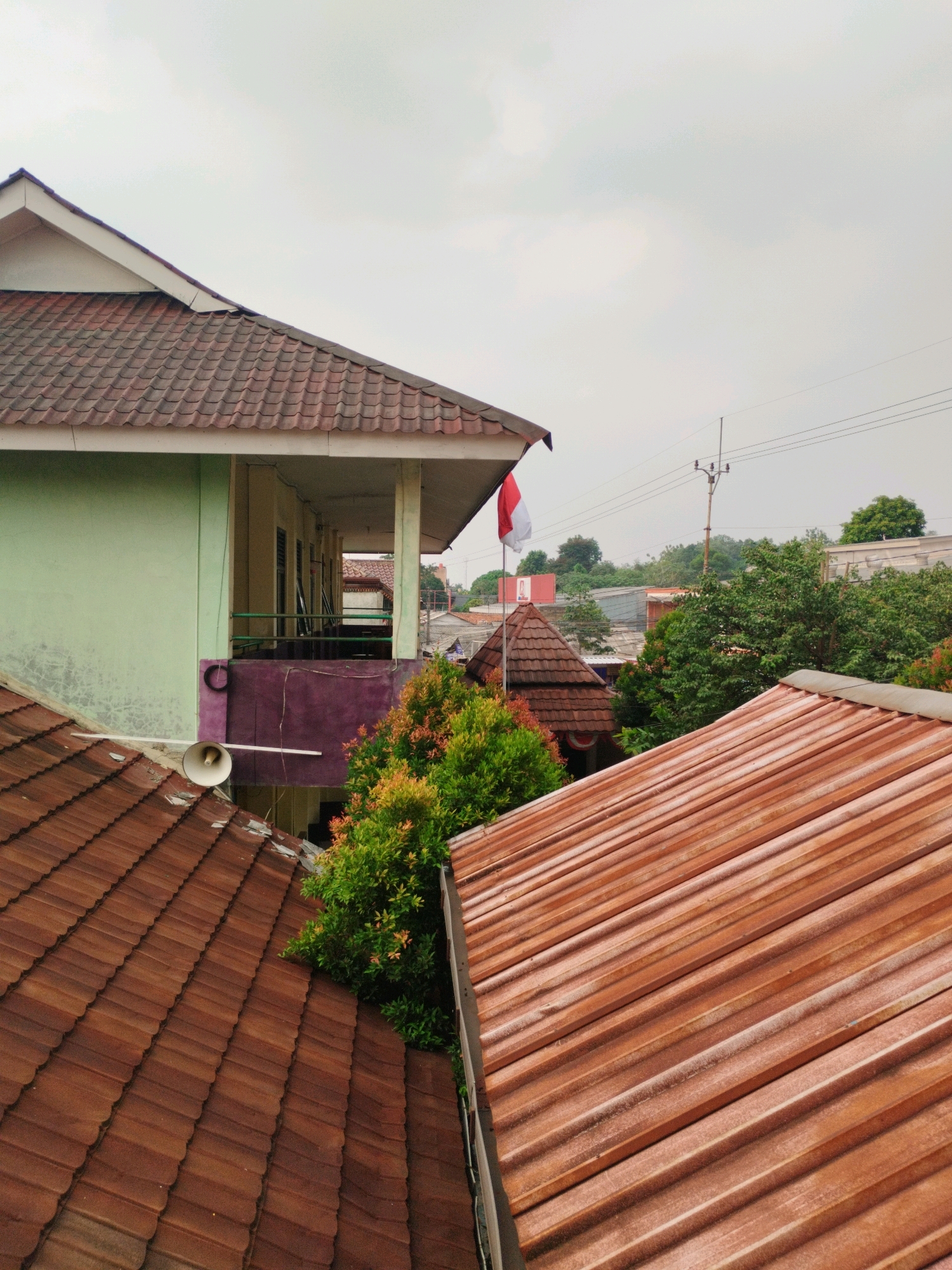 This is an eye-level, medium shot of a residential area in Depok, Indonesia, under a cloudy sky. In the foreground, two brown tiled roofs are prominent. The roof on the left is angled, while the roof on the right is flatter and has a corrugated metal appearance with streaks of dust or weathering. Behind the left roof, a light green building with a brown tiled roof and white trim is visible. A balcony with a purple railing and some architectural features protrudes from this building. A white loudspeaker is mounted on the side of the green building, above the left roof.
Further back, a small brown tiled pagoda-like structure is visible, and a red and white Indonesian flag is flying from a flagpole to its left. Beyond the buildings and vegetation, a utility pole with wires stretches across the horizon. Lush green trees and other buildings with tiled roofs fill the midground and background, suggesting a densely populated urban or suburban environment. The overall scene depicts a typical Indonesian streetscape with a focus on the architectural elements of the houses and the surrounding environment.