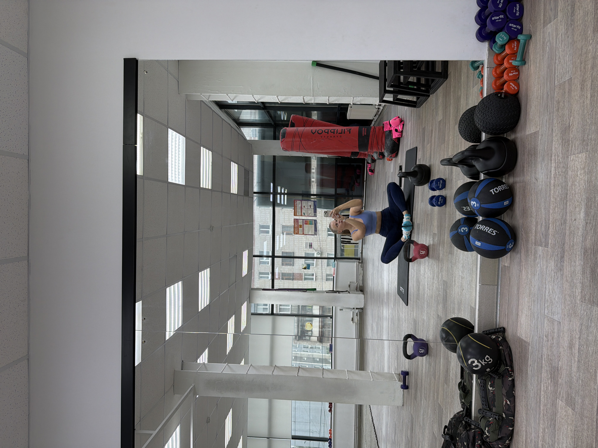 The image is an indoor scene, apparently in a gym, reflected in a large mirror. A person is visible in the mirror, sitting on a mat and taking a selfie with their smartphone. The individual is wearing a blue sports bra and dark leggings with blue athletic shoes.
Numerous pieces of gym equipment surround the person. To the right (in the reflection) are a variety of weights: dumbbells of various sizes, kettlebells, medicine balls labeled "TORRES", and sandbags. Near the top edge, small, colorful dumbbells are arranged on the floor. Behind the person, one can see a large red punching bag and pink boxing gloves. There's also a black plyometric box near the ceiling.
The room is brightly lit by fluorescent ceiling lights. Through a window, buildings can be seen, suggesting the location is in an urban area. Based on the signage and building style, this appears to be in the city of Izhevsk, Russia.