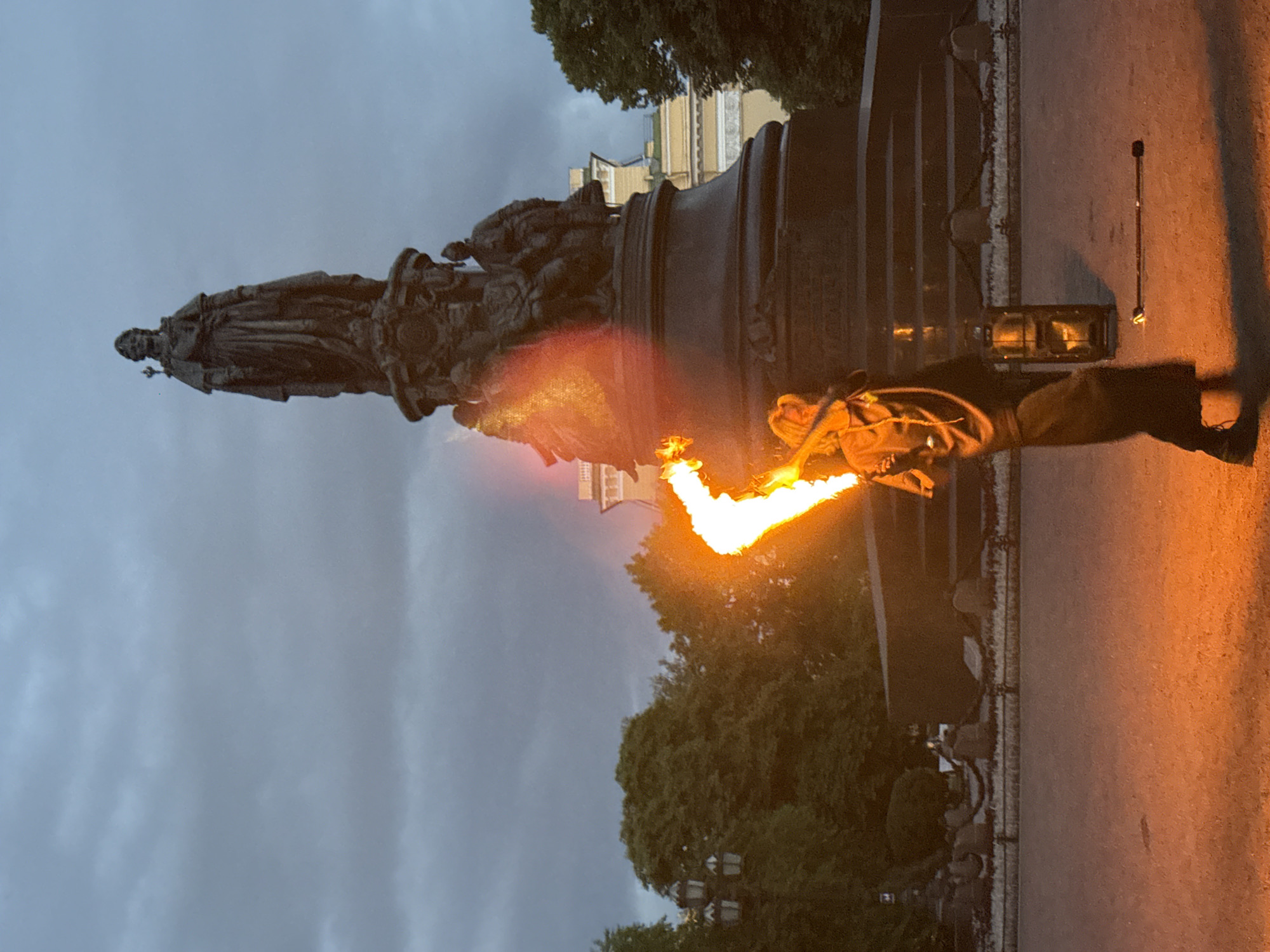 The image shows a man performing with fire in front of the Monument to Catherine the Great in Palace Square, Saint Petersburg, Russia. The sky is overcast and the lighting suggests it is dusk. The monument is a large, dark bronze sculpture of Empress Catherine II standing on a pedestal, surrounded by allegorical figures. The performer is dressed in dark clothing and is holding a flaming prop that emits a bright orange light, illuminating the immediate area. A portable lantern and a fire staff lie on the ground nearby. Trees and buildings are visible in the background, and a paved area surrounds the monument.