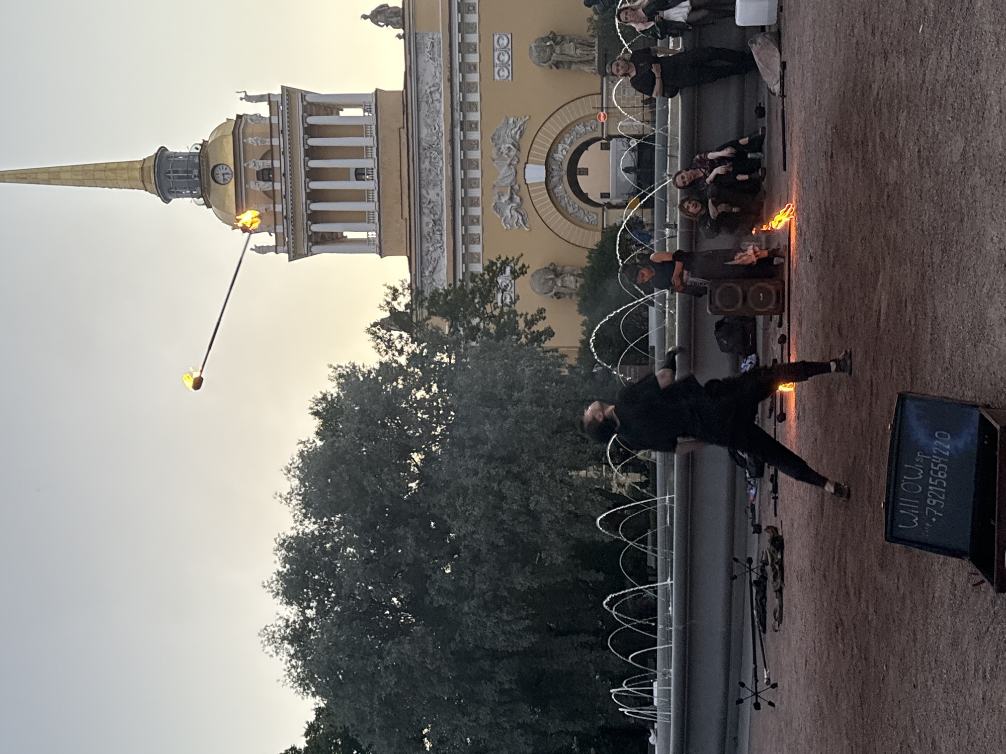 A performer is juggling a flaming prop in front of the Admiralty Building in Saint Petersburg, Russia. The performer, dressed in black, stands in the foreground, looking up at the flaming object. Several spectators are seated behind the performer, watching the show. A fountain is visible in the background, with the Admiralty Building's golden spire rising above it. The sky is overcast. The scene appears to be taking place in the early evening. A dark open case with writing on it is in the lower left of the image.