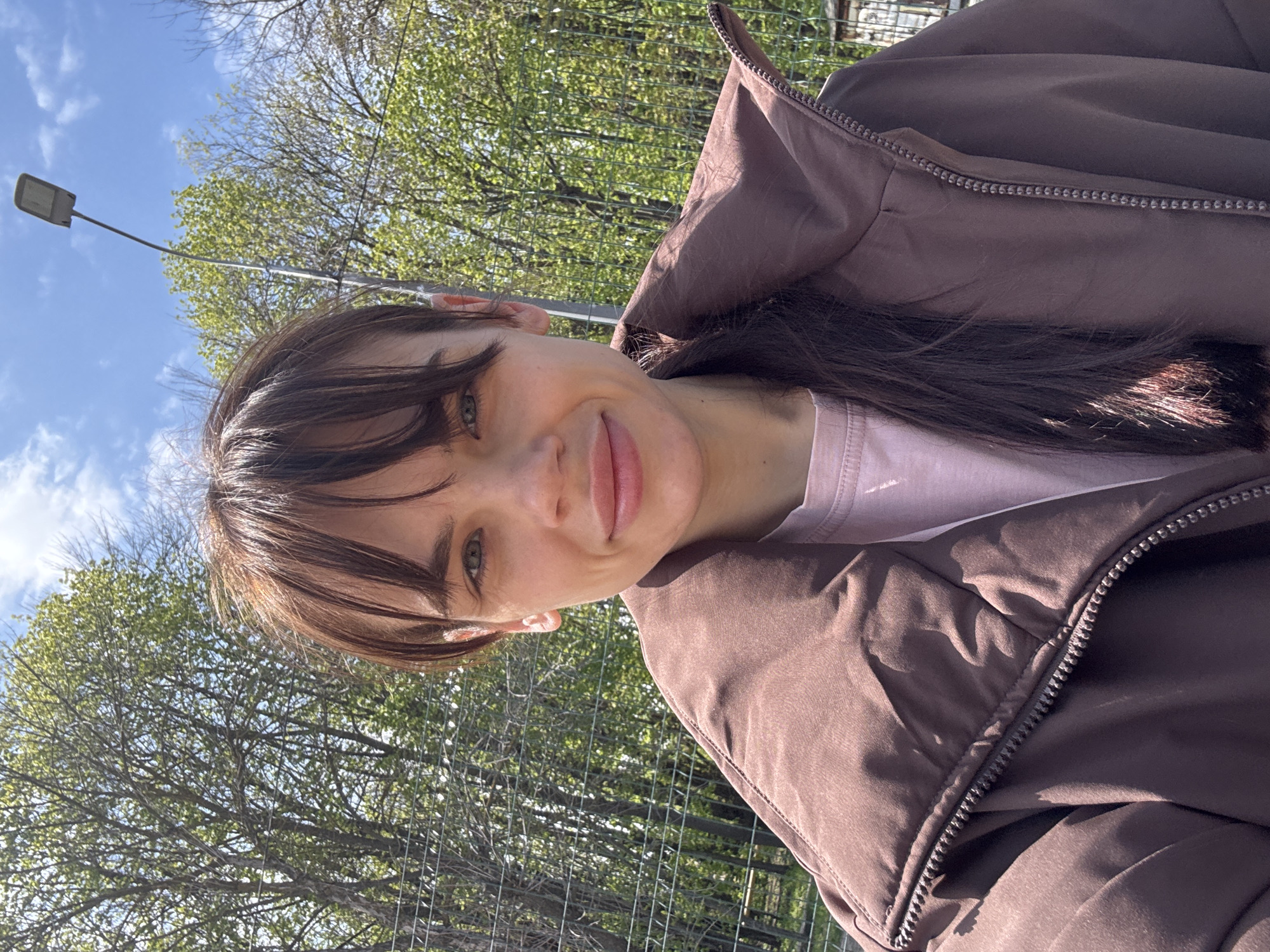 The image is a selfie of a young woman taken outdoors on a bright, sunny day, likely in Rostov-on-Don, Russia. The photo is rotated clockwise, showing the woman's head on the left and her body extending towards the right.
The central subject is the woman, who appears to be in her late teens or early twenties. She has dark brown hair styled with bangs across her forehead, and light green or hazel eyes. Her expression is calm and pleasant, with a gentle, natural smile. She is wearing a dark brown, possibly padded, jacket with a prominent silver-toned zipper, over a light pink t-shirt.
The background consists of a clear blue sky with some wispy white clouds at the top. Numerous trees with vibrant green leaves, indicating spring or early summer, are visible, along with some bare branches. A street lamp post extends into the frame from the upper left, and a metal fence or wire mesh can be seen behind the woman, suggesting a public park, recreational area, or an urban green space. The bright sunlight illuminates her face and hair, creating highlights. No visible text is present in the image.