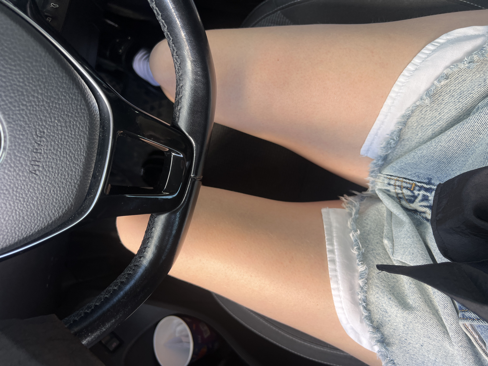 The image shows a close-up view of a person's legs in the driver's seat of a car, with a steering wheel prominently in the foreground. The person is wearing denim shorts with a white lining and a white sock. The background is dark, suggesting the interior of a car. The lighting appears to be natural, possibly from daylight, as indicated by the brightness on the legs and the steering wheel. The overall mood is casual, and the focus is on the personal space within the vehicle. No specific activity is depicted beyond occupying the driver's seat, hinting at a potential journey or simply sitting in the car. The steering wheel has the "AIRBAG" text visible on it. The context of Saint Petersburg, Russia, is provided, but there are no direct visual cues in the image to confirm this location.