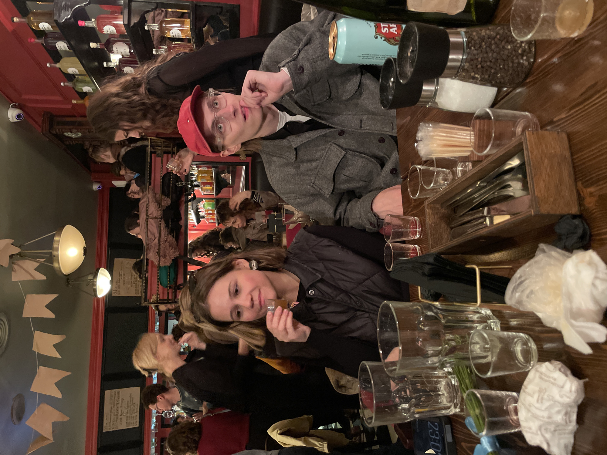 The image shows a busy interior scene, possibly a restaurant or bar. Two people are prominently positioned at a wooden table, with the table acting as a boundary between them and the general public scene. On the table are multiple clear glasses, a utensil holder containing silverware, a can of Stella Artois beer, salt and pepper grinders, toothpicks, and napkins.
The man, wearing a gray jacket and a red cap, is looking at the camera. The woman, wearing a dark jacket, is holding a shot glass and seems to be looking at something to the side. Other people are visible in the background, suggesting a crowded environment.
Flags are hanging in the background, as are the hanging ceiling lamps. Liquor bottles fill the shelf at the top of the image.
The city and specific location are not discernible from the image.