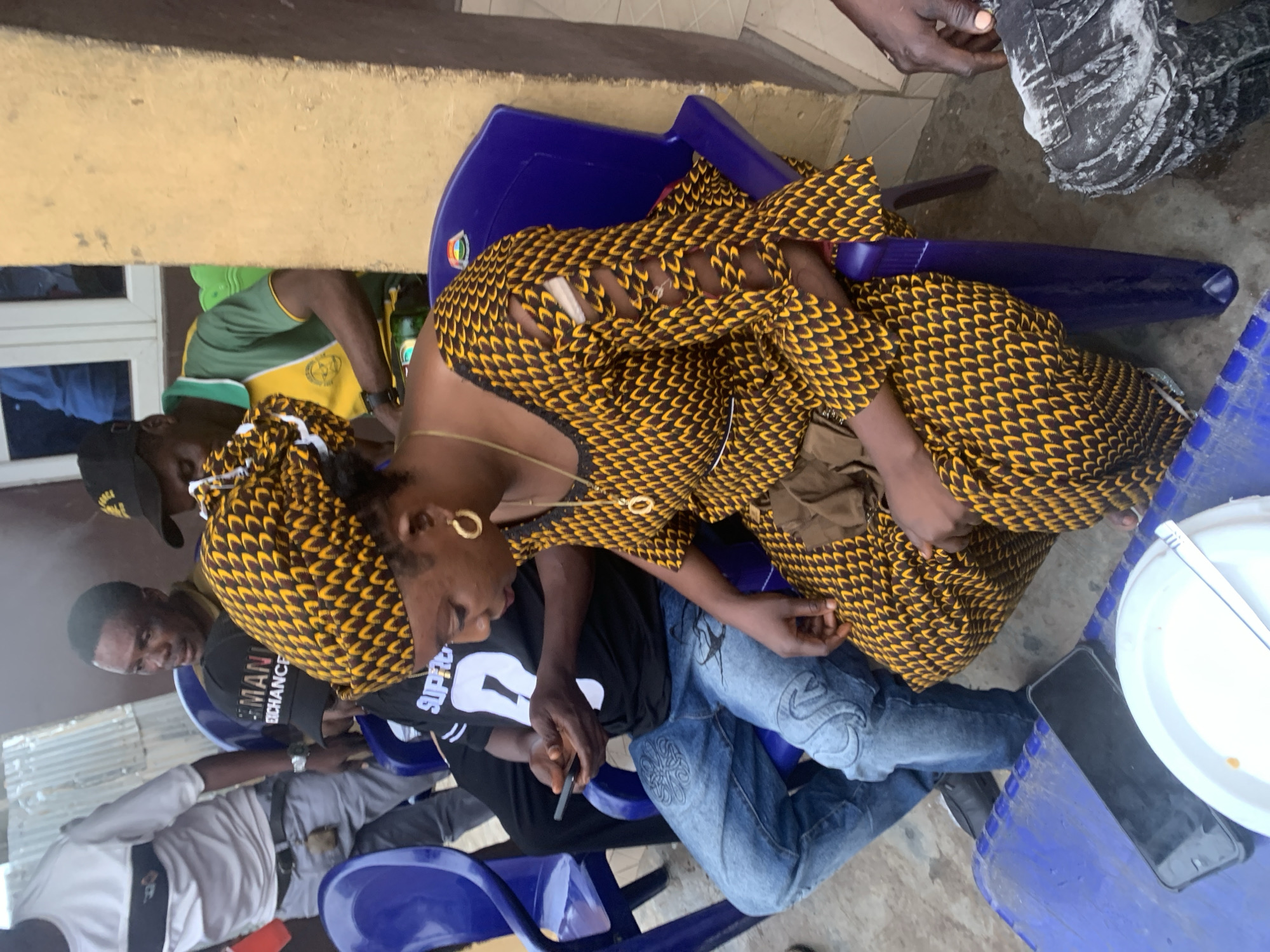 In Shagamu, Nigeria, a group of people are gathered outdoors. A woman in the foreground wears a vibrant yellow and black patterned dress and headscarf, with a gold necklace and hoop earrings. She is seated on a blue plastic chair, holding a brown pouch. Beside her, a man in a black t-shirt with a white number '8' and "SUPREME" text, and light blue jeans with decorative embroidery, is also seated and looking at his mobile phone. To his left, another man wears a black cap and a yellow and green t-shirt, also seated. In the background, a man in a white t-shirt and grey trousers stands. The environment appears to be a casual outdoor setting, possibly near a building with yellow walls and a window. Blue plastic tables are visible in the foreground, one with a white plate and a phone. The ground is rough, possibly concrete or dirt.
