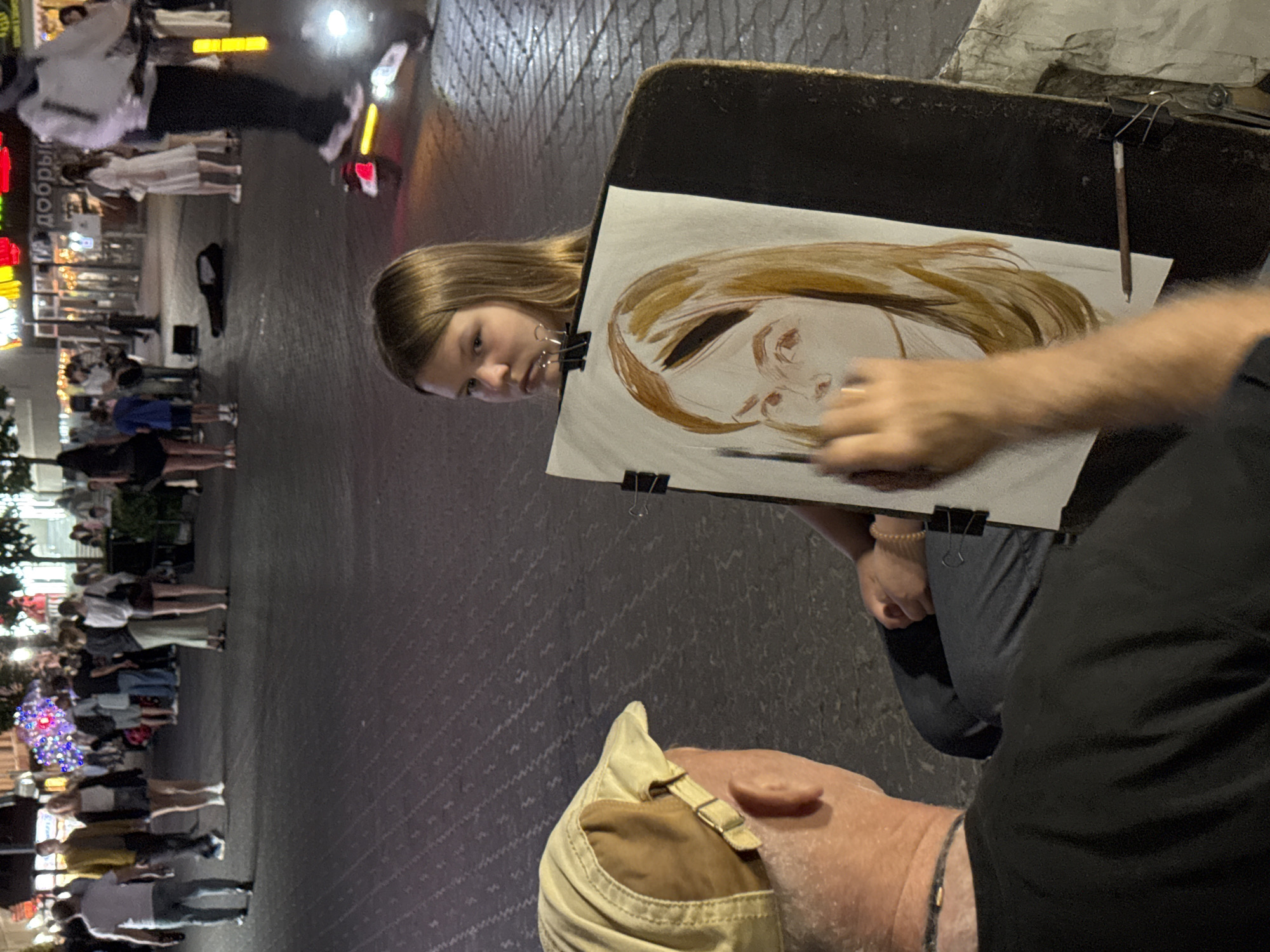In Kazan, Russia, on a city street at night, a street artist is painting a portrait of a young girl who is posing for him. The artist, an older man wearing a beige baseball cap and a black t-shirt, is in the foreground, facing away from the camera, with his left arm extended as he paints on a canvas propped up on an easel. The young girl, with shoulder-length brown hair, is looking to her right with a neutral expression. The portrait on the canvas is of her face, with unfinished features and colored hair. The background is blurred, showing other people walking on the street, some buildings with lights, and a person on an electric scooter. The street appears to be paved with cobblestones.