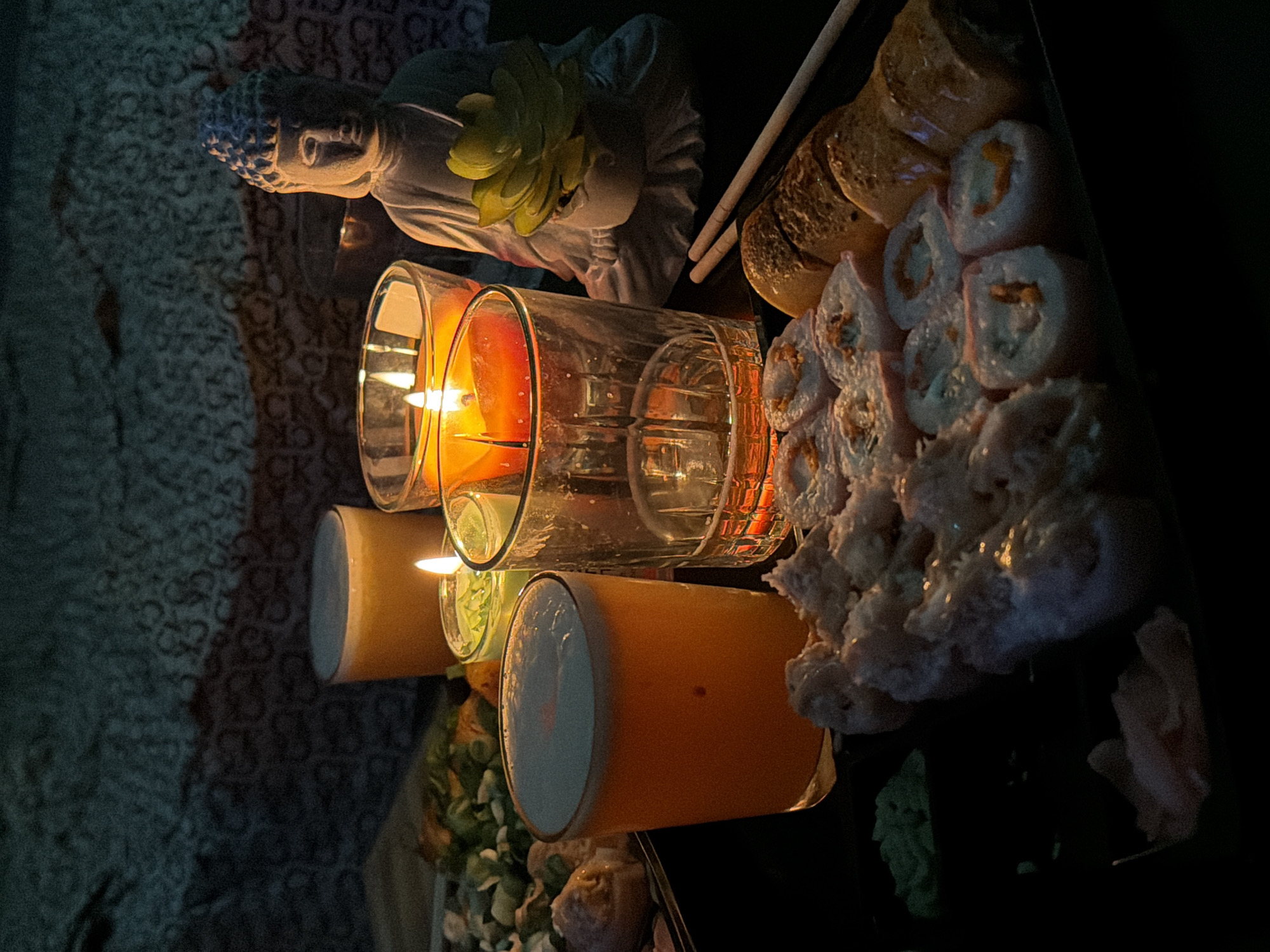The image captures a cozy, dimly lit indoor setting, likely at night, in Varna, Bulgaria. A tranquil scene unfolds on a dark, textured surface, possibly a tablecloth or blanket, which features a subtle, repeating pattern of letters like "CKCKCK...".
On the left, a serene light-colored Buddha statue, possibly made of stone or ceramic, is visible. A small green succulent or artificial plant is positioned behind its head, adding a touch of nature.
The center of the frame is illuminated by the warm glow of candlelight. Two candles burn within glass holders; one is prominently displayed in an orange-tinted glass, casting a rich, warm light, while another smaller candle sits in a clear glass. Adjacent to the candles are two tall drinking glasses. One holds a vibrant orange or yellow beverage topped with a frothy layer, and the other contains a lighter, possibly clear, liquid.
To the right, a selection of sushi is arranged on a dark platter. Various rolls are present, some featuring white rice with a light orange or pink filling, and others appearing to be different types of sushi or rolls. A pair of chopsticks rests nearby, indicating an ongoing or impending meal. Accompanying the sushi are small dollops of green wasabi and slices of pink pickled ginger. The overall atmosphere is one of calm and relaxation, suggesting a pleasant dining experience.