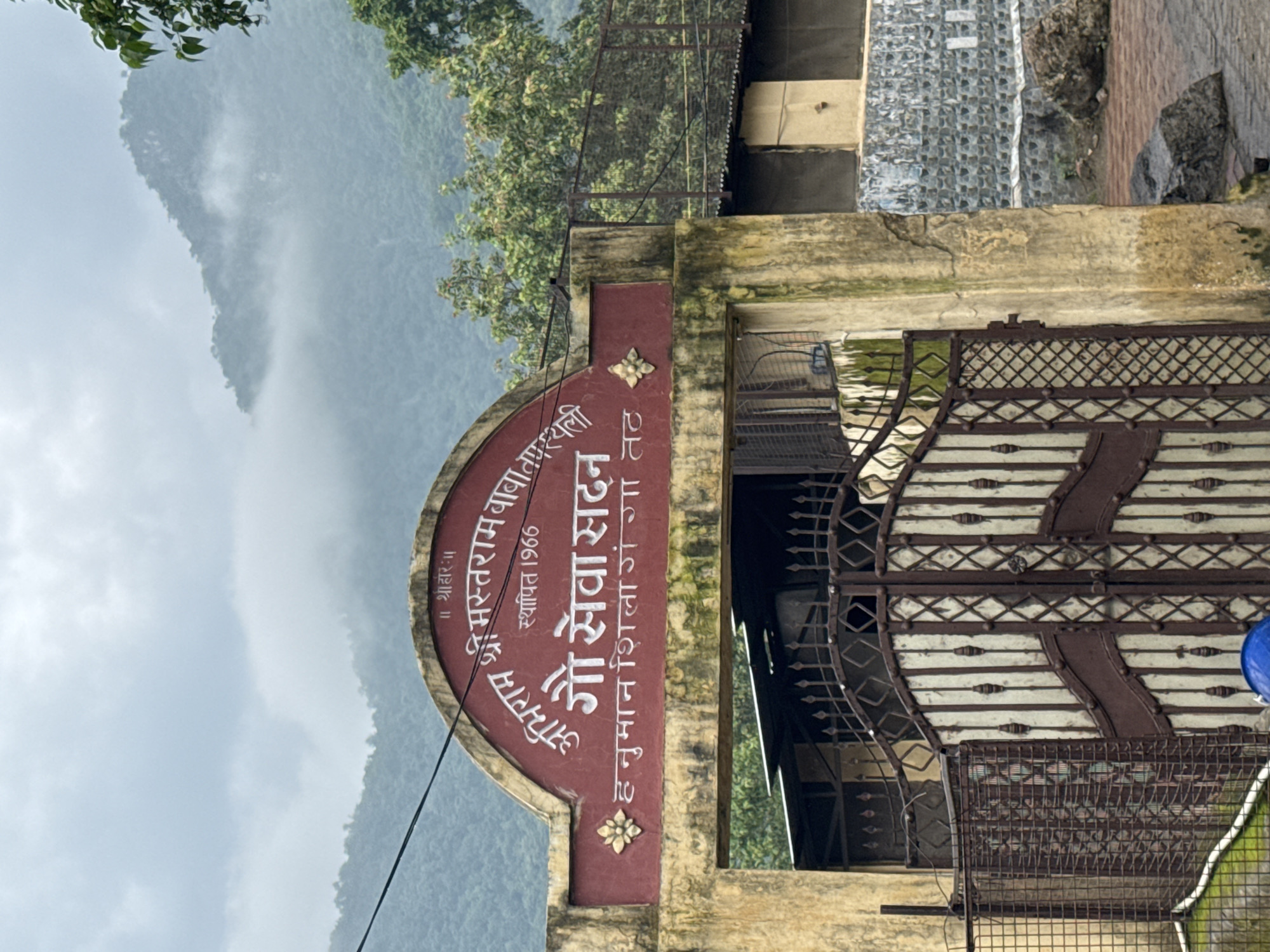 The image shows the entrance to "Gau Seva Sadan" (Cow Service Center) in Rishikesh, India. The building's facade is painted a weathered, pale yellow with a prominent maroon archway above the entrance. Inscribed within the arch in white lettering are Hindi words. At the top, it reads "॥ श्रीहारः ॥" and below that, "श्री मस्तराम बाबा की". To the right of this, "स्थापित 1966" (Established 1966) is visible. The central part of the arch displays "गौ सेवा सदन" (Gau Seva Sadan) in large white letters, and below that, "हनुमान शिला गंगा तट" (Hanuman Shila Ganga Tat). A decorative golden motif, resembling a flower, is present on the left side of the arch.
The entrance is guarded by a tall, ornate gate made of brown metal with cream-colored vertical panels. To the left of the main gate is a section of wire mesh fencing. Beyond the gate, a dark, covered structure is partially visible. The background features lush green trees and the misty outline of mountains, suggesting a natural, possibly humid, environment. The ground in front of the gate is paved with red bricks and there are a couple of large rocks on the right side of the image. A blue object, possibly a helmet, is visible in the lower center of the frame.