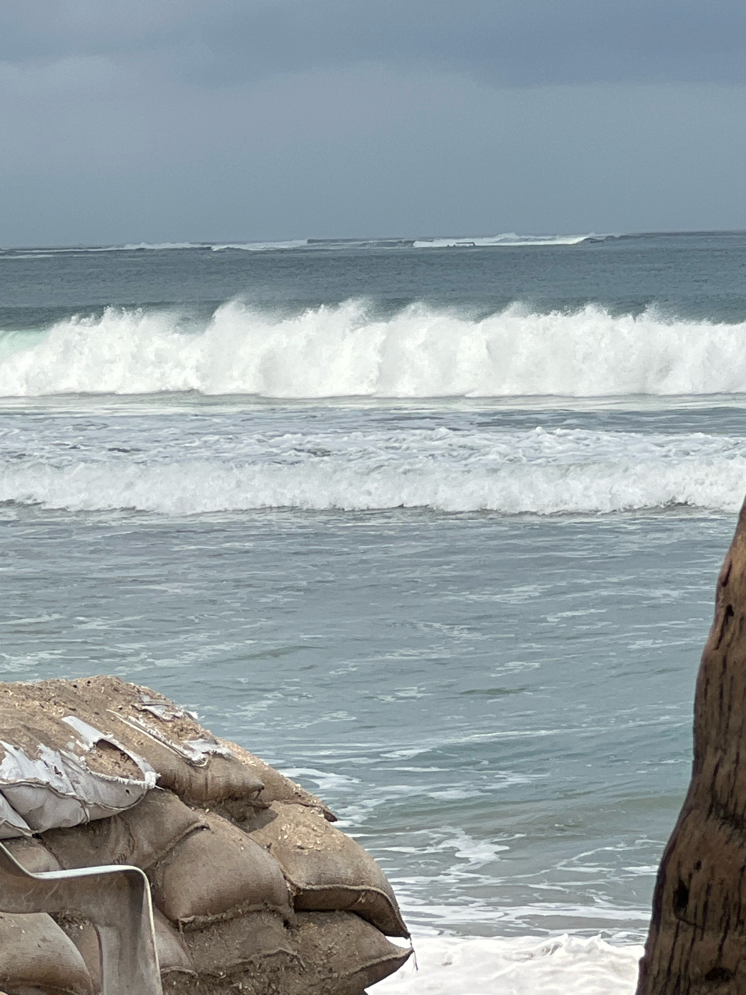 In Kuta, Indonesia, a beach scene unfolds under an overcast sky. The ocean is active with several lines of waves. The closest waves to the shore are smaller and foamy, while further out, larger, powerful waves are breaking, forming a white, frothy crest. In the foreground, a wall of sandbags, some showing signs of wear and tear, is piled up on the beach. A metal scoop is visible amongst the sandbags. To the right, the textured surface of a large piece of driftwood frames the scene. The overall impression is one of a protected coastline preparing for or enduring rough seas.