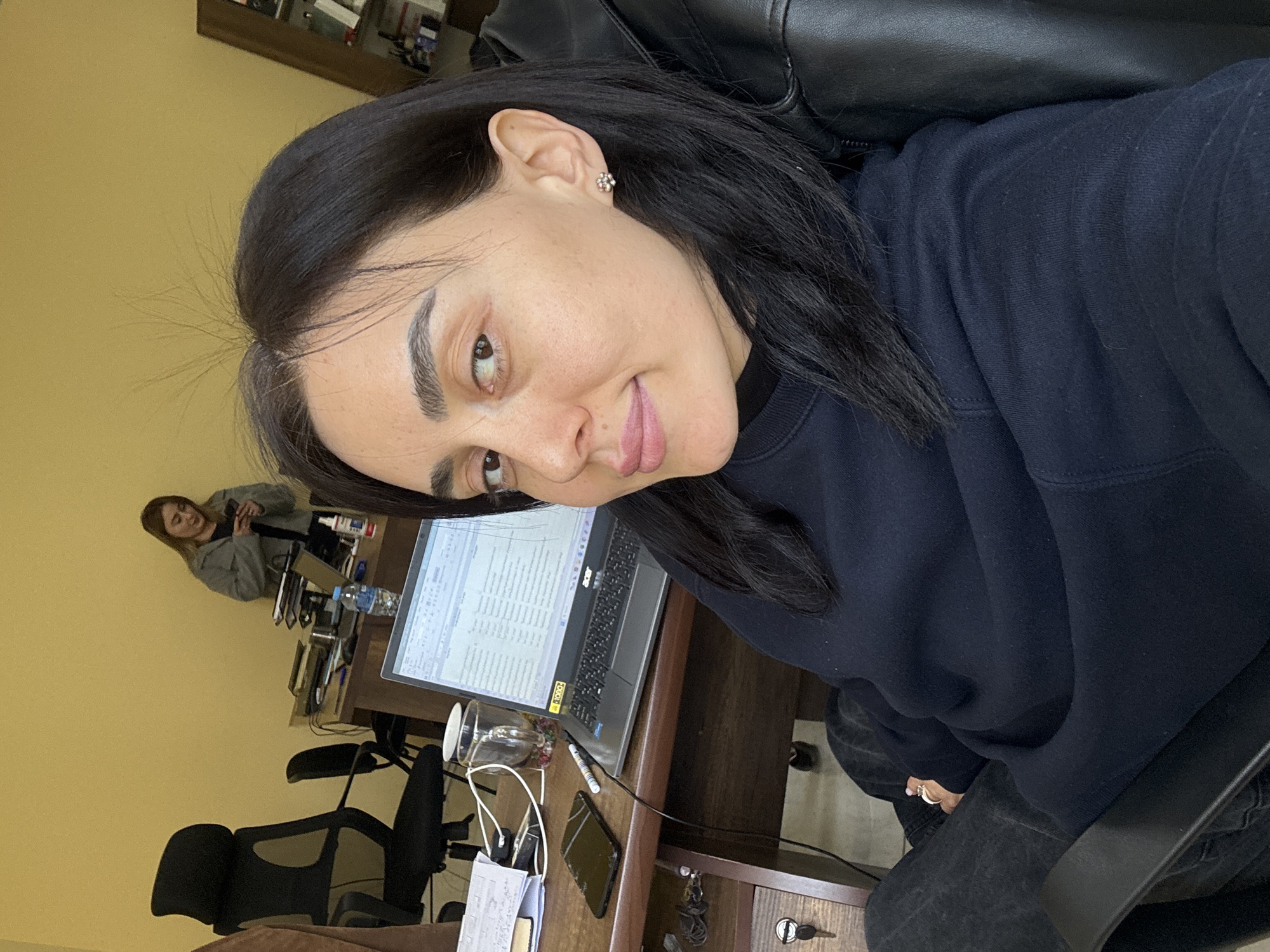 A woman with dark hair and a dark sweatshirt is sitting in a black chair at a desk. A laptop is open on the desk, along with papers, a phone, and a glass. Another woman is visible in the background, sitting at another desk. The room has yellow walls. The location is likely an office setting in Tbilisi, Georgia.