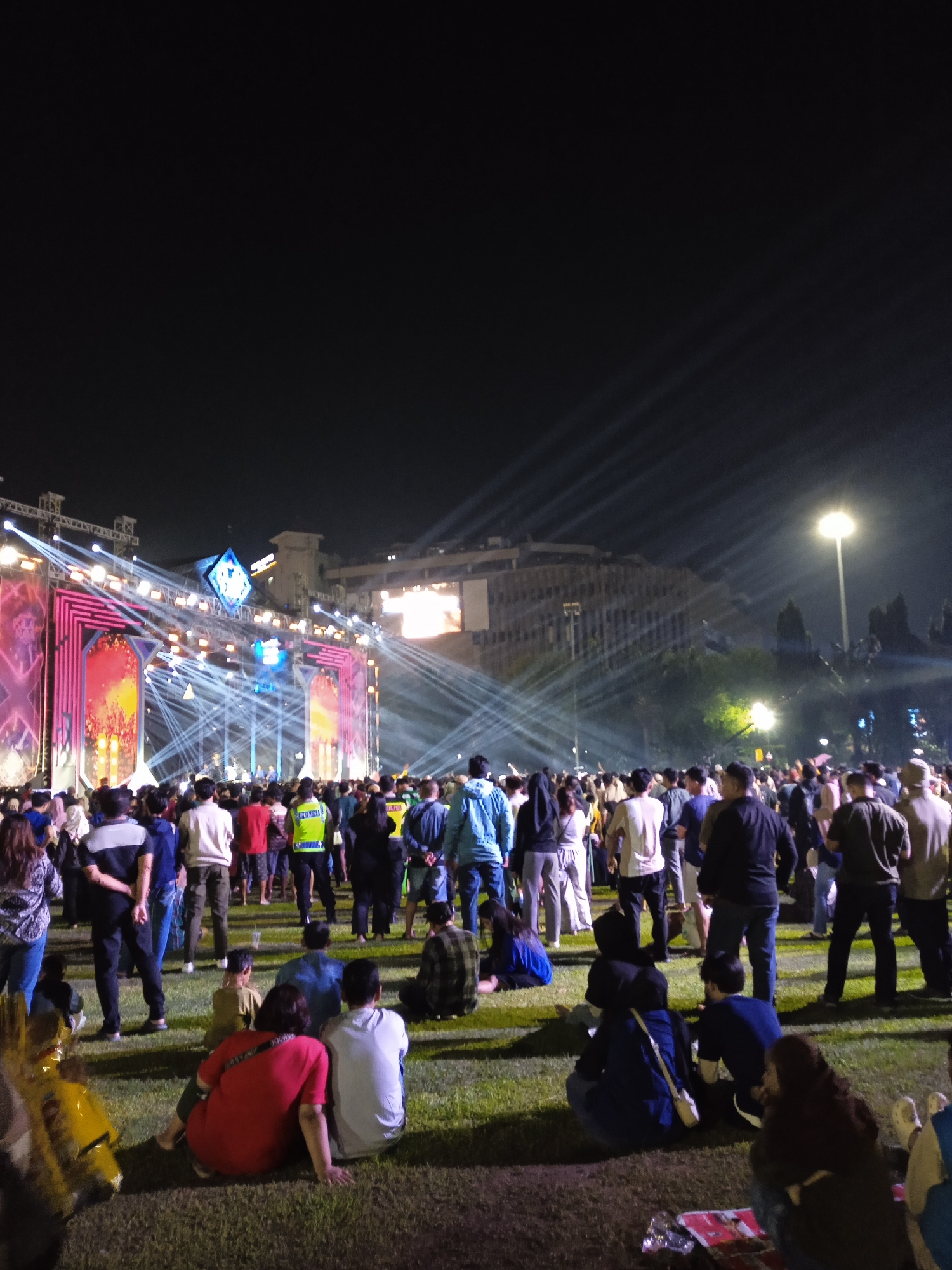 At night in Semarang, Indonesia, a large crowd gathers on a grassy field facing an illuminated stage. Light beams shoot into the sky, and a tall building is visible in the background. People stand and sit on the grass, and a uniformed police officer is visible in the crowd.