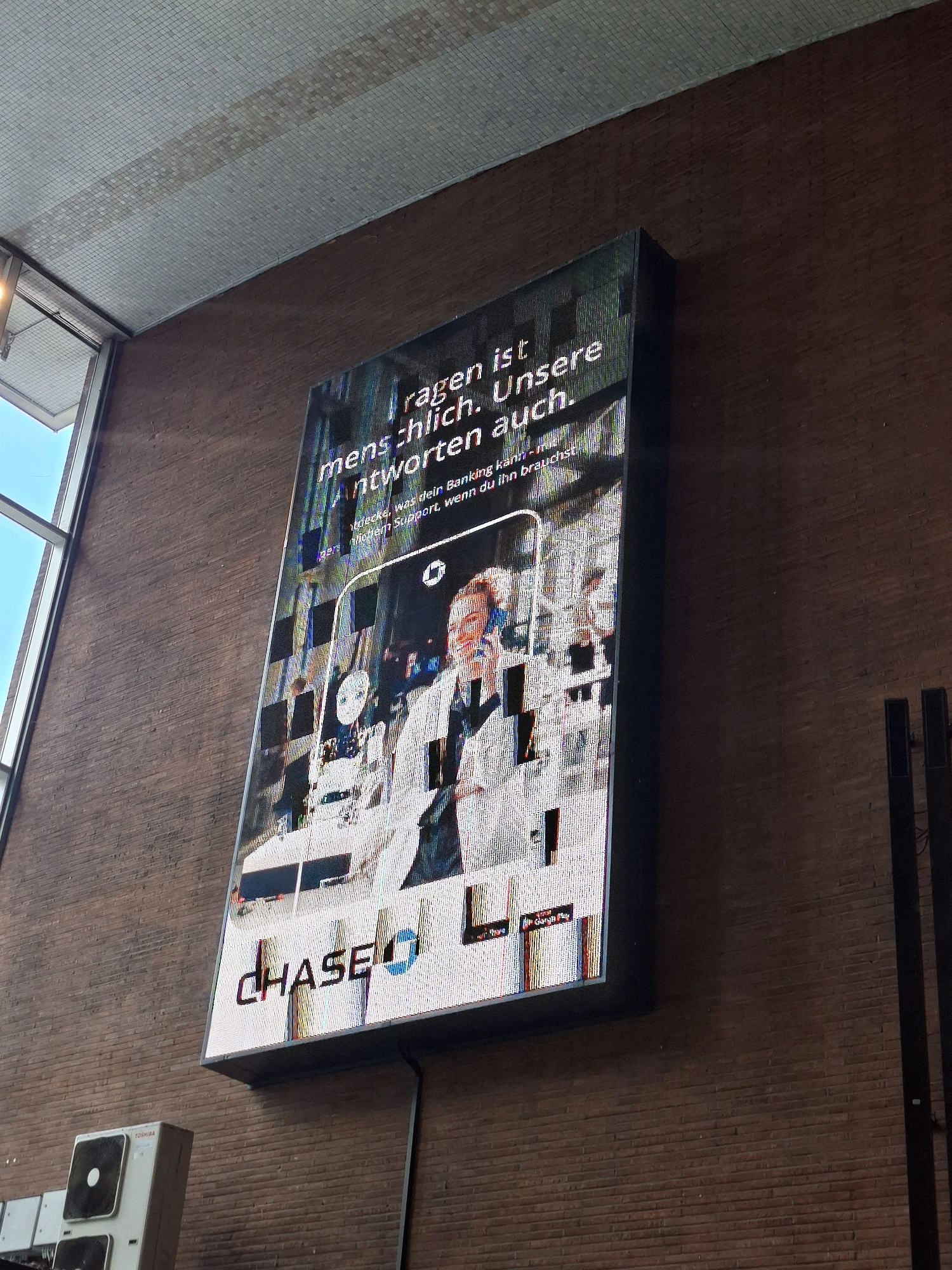 An outdoor digital billboard is mounted on a brick wall, displaying an advertisement for Chase Bank. The billboard shows a smiling man in a suit holding a smartphone, with text in German reading "Fragen ist menschlich. Unsere Antworten auch." which translates to "Asking is human. Our answers too." Below this text, smaller print reads "Entdecke, was dein Banking kann – mit modernem Support, wenn du ihn brauchst." or "Discover what your banking can do - with modern support when you need it." The Chase logo is prominently displayed at the bottom of the billboard. The billboard is slightly tilted and shows some digital distortion. In the background, a window with a blue sky is visible to the left, and two black vertical speakers are mounted on the wall to the right. Below the billboard, an air conditioning unit with a Toshiba logo is visible on the brick wall. The scene appears to be in Cologne, Germany, indicated by the presence of a Chase Bank advertisement, as Chase Bank operates internationally.