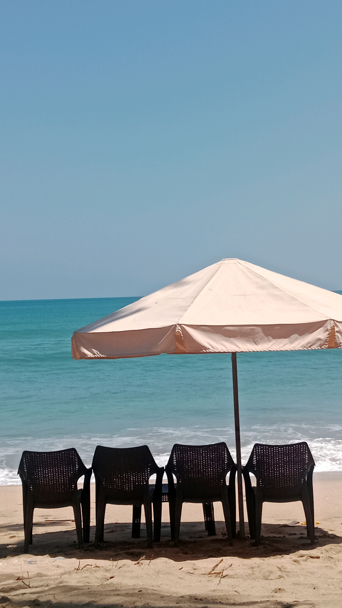 Four dark plastic chairs are positioned on a sandy beach in front of a light-colored beach umbrella. The umbrella is supported by a central pole and casts a shadow on the sand. Behind the chairs and umbrella, the turquoise ocean meets the horizon under a clear blue sky. The scene appears to be in Kuta, Indonesia, a popular tourist destination known for its beaches and vibrant atmosphere. There are no people or animals visible in the image. The overall mood is peaceful and serene, suggesting a quiet moment on the beach.