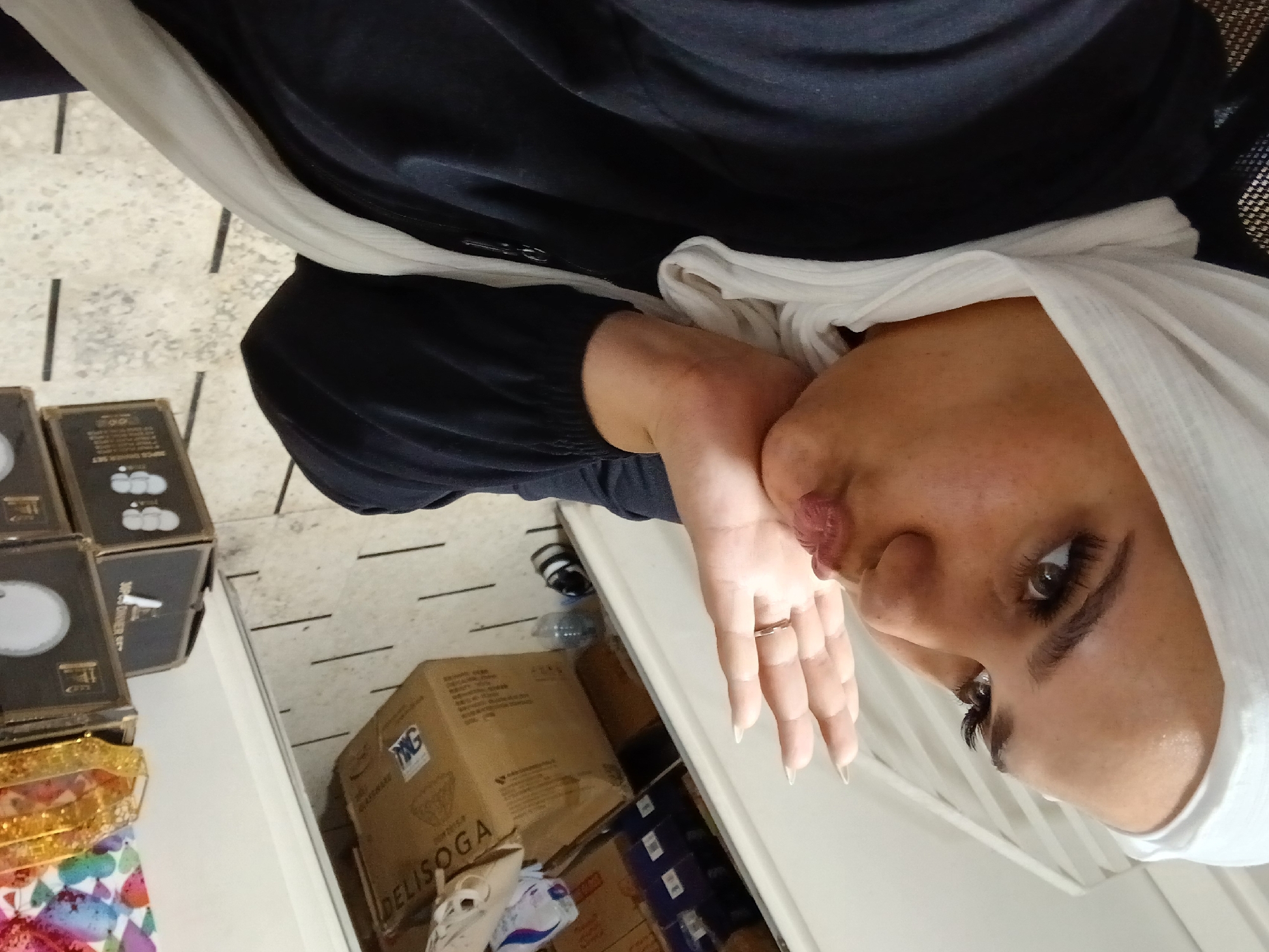 A woman wearing a white hijab and a dark blue robe is blowing a kiss towards the camera. She has long, dark eyelashes and a ring on her right ring finger. Her left hand is raised and open, with her palm facing the camera. The background appears to be a room with boxes stacked on shelves. One box has the text "DELISOGGA" printed on it. The floor is tiled with a light-colored stone and has black lines. The image is taken from a high angle, with the woman's face dominating the foreground.