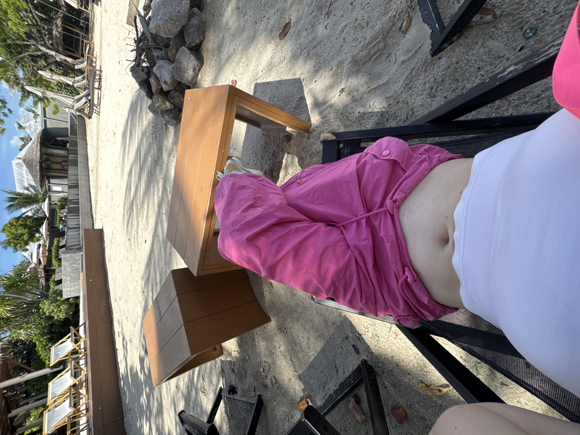 A person is lounging on a black chair on a sandy beach, wearing a pink shirt and white shorts. A wooden table is positioned next to the chair, and a pile of rocks is visible behind it. In the background, there are trees, umbrellas, lounge chairs, and buildings with thatched roofs, suggesting a resort or hotel setting. The sun is casting shadows on the sand, indicating it is daytime. The scene appears to be a tranquil vacation spot.