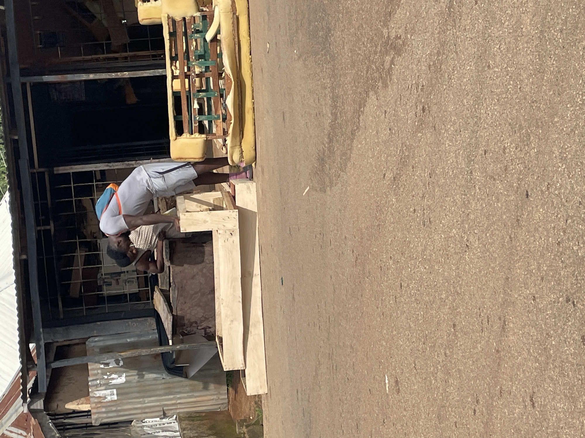 In Jos, Nigeria, two men are visible in front of a weathered storefront, its corrugated metal and wooden construction suggesting a workshop or informal business. The man in the foreground, wearing a grey t-shirt, grey shorts, and pink shoes, leans over a wooden frame. He has a blue and orange backpack slung over his left shoulder. Behind him, another man, wearing a dark tank top, is also engaged with the wooden structure. Nearby, a dilapidated yellow sofa frame rests on the asphalt road, its upholstery partially removed, revealing a wooden and metal substructure. The ground is paved with asphalt, showing signs of wear and scattered debris. The overall impression is of a bustling urban environment, likely a furniture repair or manufacturing area.