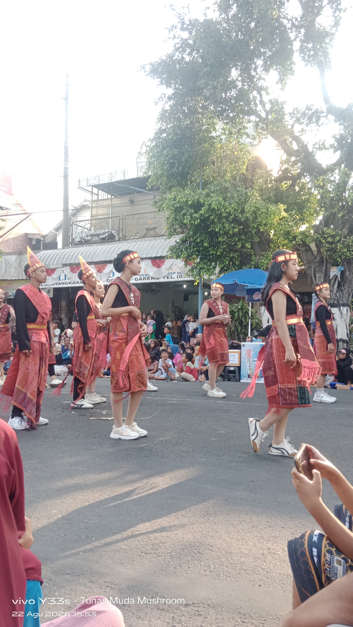 In the city of Genengan, Indonesia, a group of young people in traditional attire participate in a street parade. The individuals, likely performing a cultural dance, are dressed in vibrant red and gold outfits featuring intricate patterns. They wear matching headbands and have their hair styled. Some carry props or accessories that are part of their performance.
The parade takes place on a paved street, with spectators seated on the side, watching the procession. Behind the participants, buildings and trees form the urban backdrop, with the sun casting long shadows, indicating it might be late afternoon. The overall atmosphere appears festive and community-oriented. The timestamp at the bottom of the image, "22 Agu 2026 15:53," suggests the event was captured on August 22, 2026, at 3:53 PM.