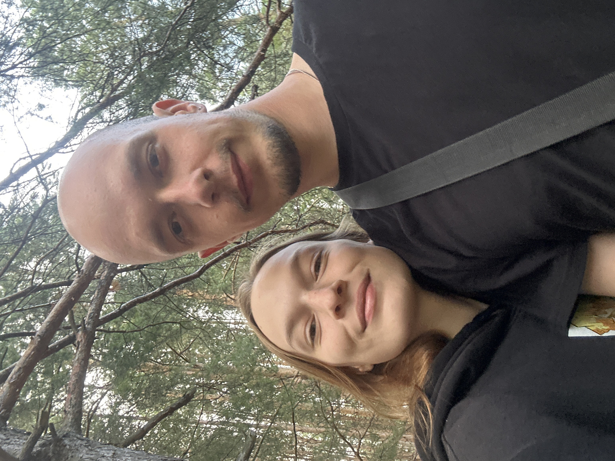 A selfie shows a young Caucasian couple posing for a photo in a forest. The man, who is bald with a goatee, is on the right side of the frame. He is wearing a black t-shirt and a grey strap across his chest. The woman, who has blonde hair, is on the left side of the frame. She is wearing a black hoodie. They are both smiling at the camera. The background is filled with trees and foliage, suggesting a natural, outdoor environment. The lighting indicates it is daytime, possibly overcast. The image appears to have been taken in the city of Chaikovsky, Russia, as indicated by the context.