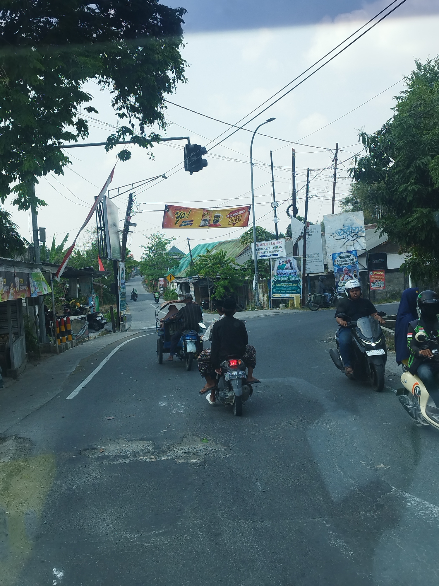 The image depicts a street scene in Baureno, Indonesia, on an overcast day. Several motorcycles and a three-wheeled vehicle are on a paved road with some visible potholes and markings. In the foreground, a person on a motorcycle, wearing a white helmet and dark clothing, is riding forward. To their right, another person on a scooter with a blue and green covering is also moving. Further ahead, a three-wheeled vehicle carries two individuals. On the left side of the road, there are various structures, including shops with awnings, signs, and what appear to be advertisements. A traffic light is visible overhead, and numerous electrical wires crisscross the sky. Green trees and foliage line the sides of the street. The background shows more buildings and a slightly uphill road.