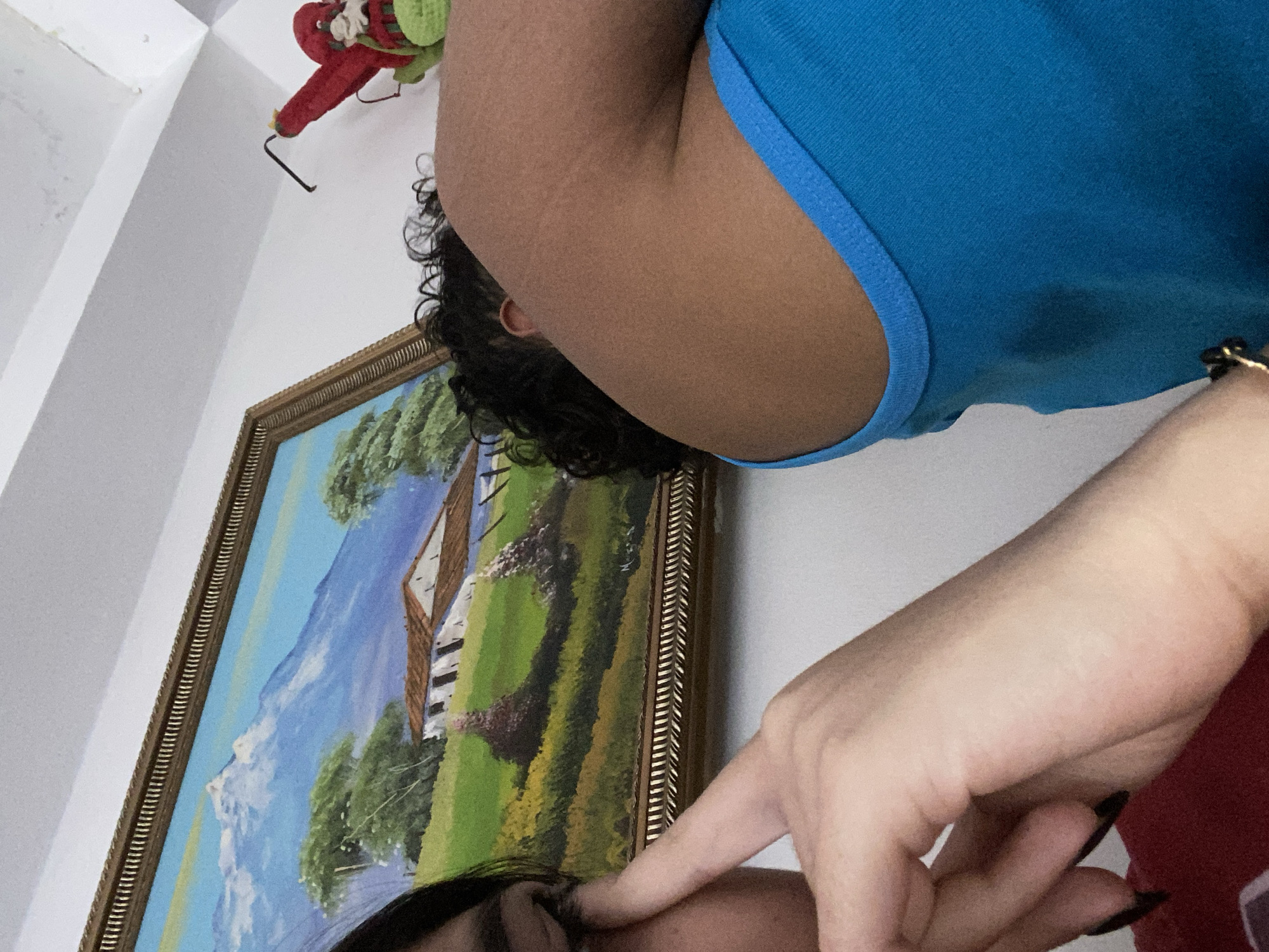A photograph, rotated counterclockwise, depicts a close-up view of a person in a blue tank top. A portion of their head is visible, featuring dark, curly hair. Their hand, adorned with black nail polish, is positioned near their face.
Behind the person is a framed landscape painting, featuring a mountain scene with a house, trees, and a blue sky. In the upper left corner of the image, there is a small decorative figure hanging on the wall.
There is no information to suggest this image was taken in Cotoperíz, Venezuela.