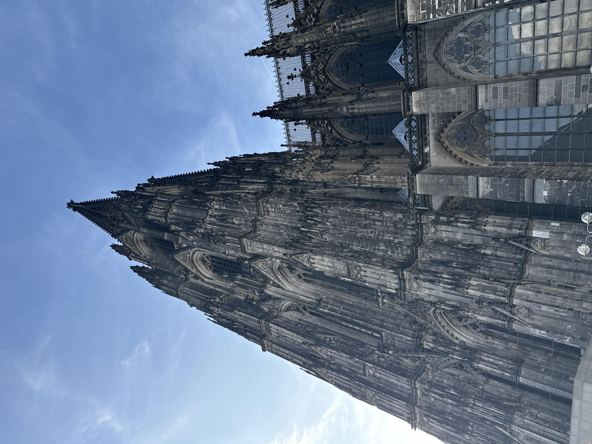 This is a low-angle, full-shot photograph of the Cologne Cathedral, a Roman Catholic cathedral in Cologne, Germany. The cathedral's towering spires and intricate Gothic architecture dominate the frame, reaching towards a clear blue sky with scattered wispy clouds. The stone facade of the cathedral is dark and detailed, showcasing numerous windows, arches, and decorative elements. The sun is not directly visible, but its light illuminates the building and creates subtle shadows. The scene is devoid of any people or animals. The environment is urban, with the sky and the cathedral being the primary visual elements.