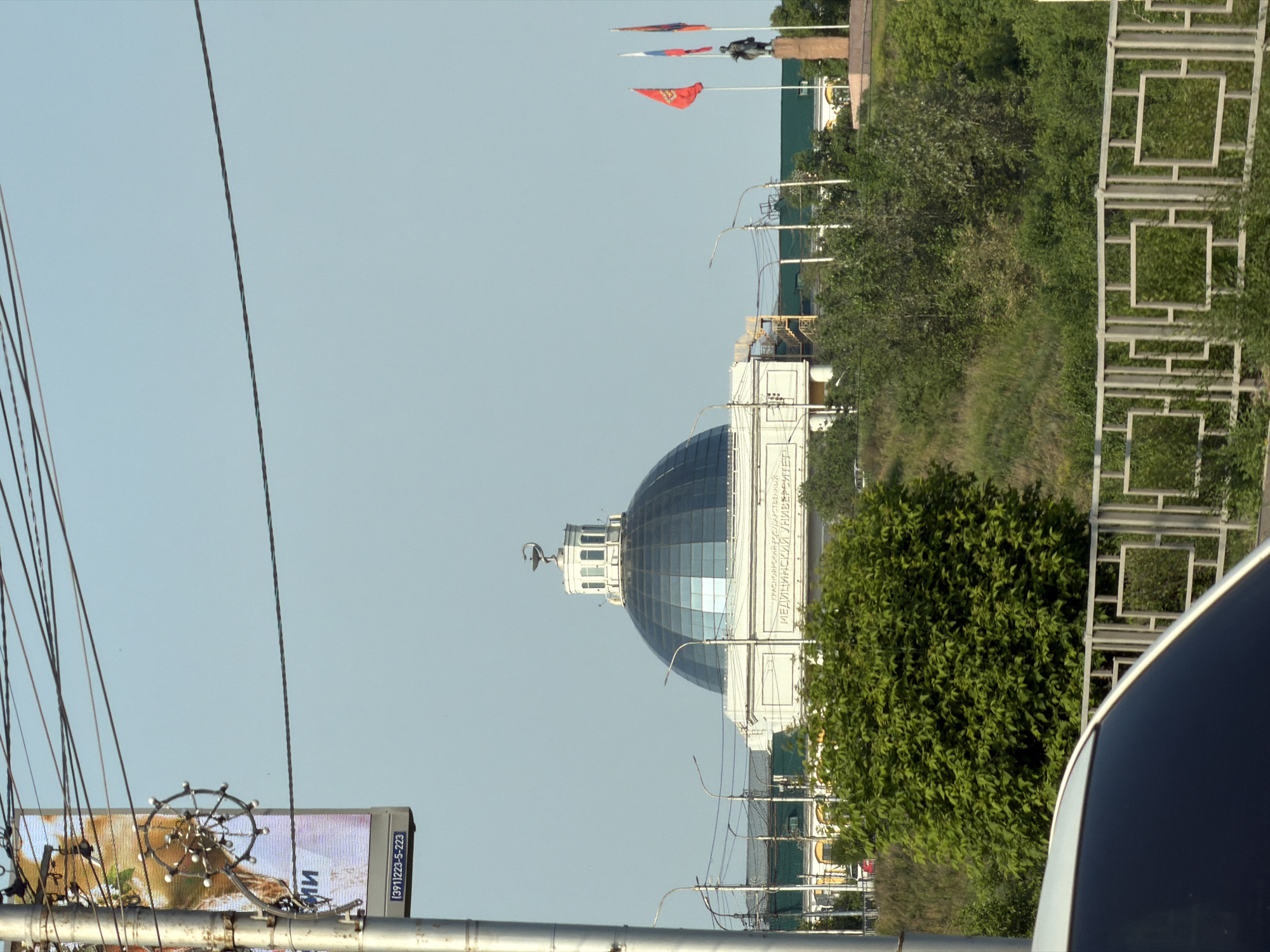 The image captures a street-level view in Krasnoyarsk, Russia, focusing on a building with a prominent blue dome and a sign reading "КРАСНОЯРСКИЙ ГОСУДАРСТВЕННЫЙ МЕДИЦИНСКИЙ УНИВЕРСИТЕТ" (Krasnoyarsk State Medical University). A statue stands to the right of the building, accompanied by three flags. In the foreground, a white car's hood is partially visible, along with a decorative metal fence. Overhead, a dense network of power lines crisscrosses the sky, obscuring parts of the view. A street pole with a sign displaying a phone number is on the left side of the frame. The scene is framed by lush green trees and vegetation, suggesting a daytime setting.