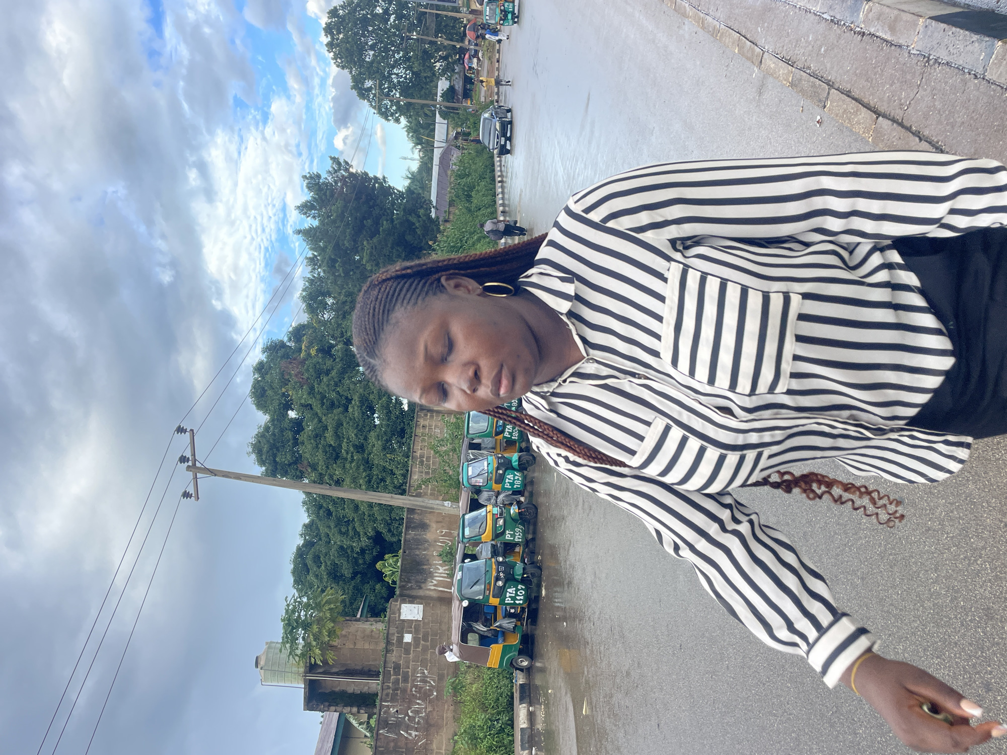 In Jos, Nigeria, a young woman with dark braided hair walks along a wet asphalt road on an overcast day. She wears a black and white striped button-down shirt with a pocket and black pants. Her eyes are closed, and her gaze is directed downwards. In the background, a row of green and yellow auto-rickshaws are parked on the side of the road, some displaying license plates. A brick wall with graffiti runs along the roadside, and beyond it, lush green trees form a dense backdrop. A wooden utility pole with electrical wires stands near the wall. Further down the road, other vehicles and pedestrians are visible. The environment appears to be a typical urban street scene in Nigeria.