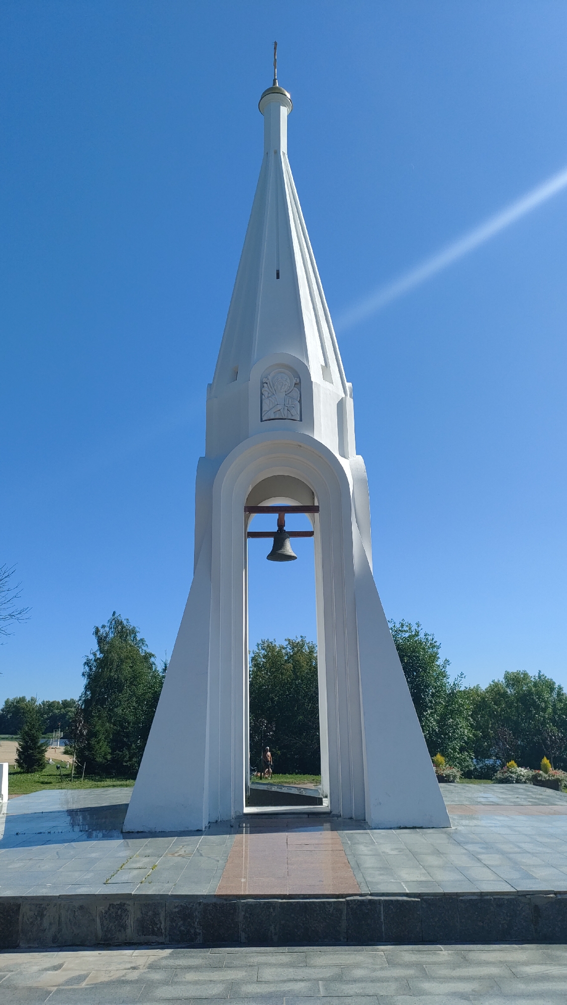 The image shows a white bell tower with a spire, set against a clear blue sky. The bell tower has an arched opening in the center, through which a bell is visible. There is a relief sculpture above the arch. The tower is situated on a paved area with trees and a grassy area in the background. In the background, a person can be seen walking with a dog. The bell tower appears to be located in Yaroslavl, Russia, specifically at the Church of Elijah the Prophet.