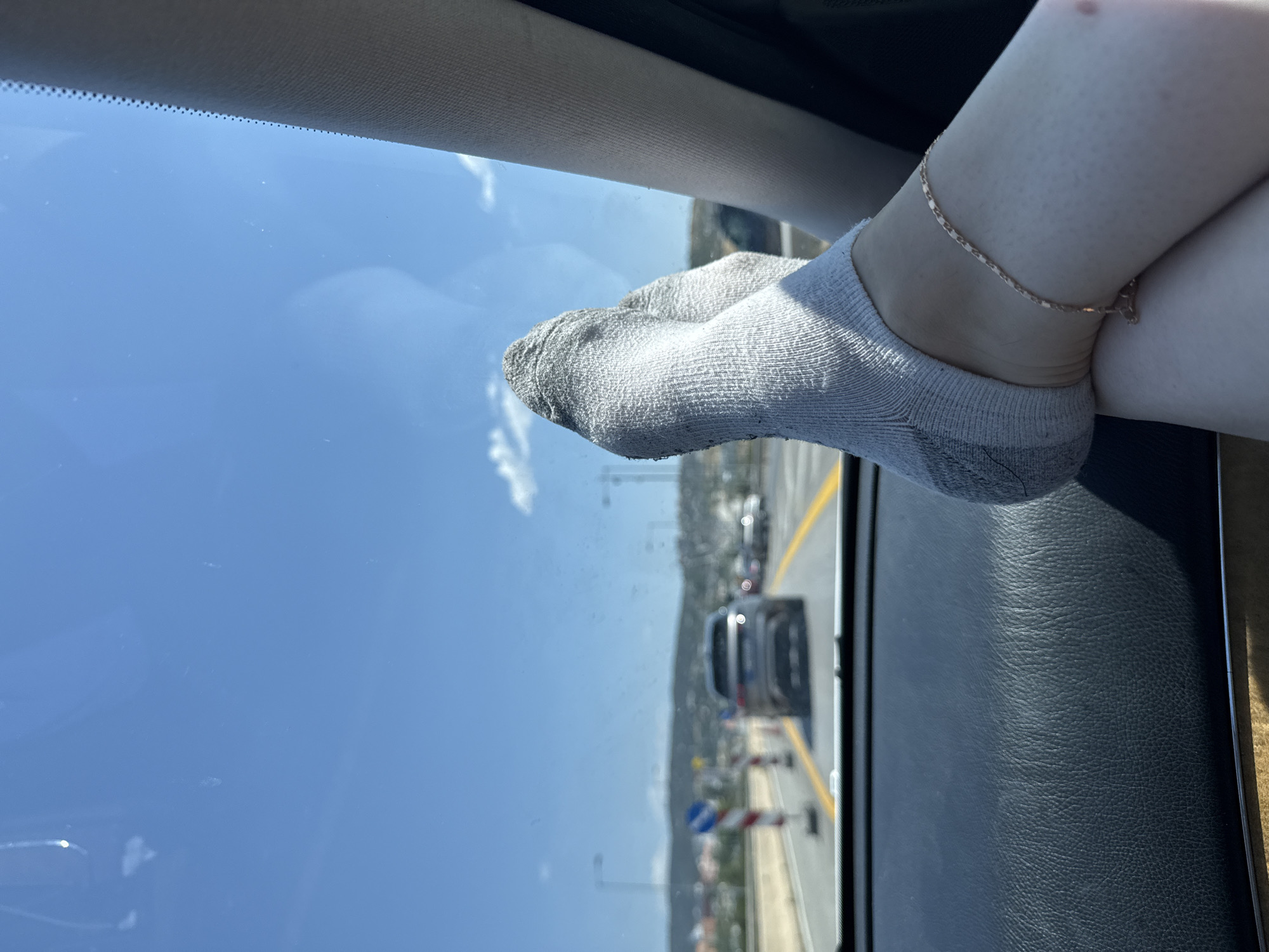 The image shows a close-up view of a person's legs and feet, wearing light grey ankle socks and a thin gold chain anklet, resting on the dashboard of a car. The perspective is from inside the vehicle, looking forward through the windshield. Outside the car, a highway is visible with other vehicles, traffic cones, and road markings. The sky is clear and blue with some scattered clouds. The dashboard is made of black, textured material. The location is identified as Efkarpia Municipal Unit, Greece.