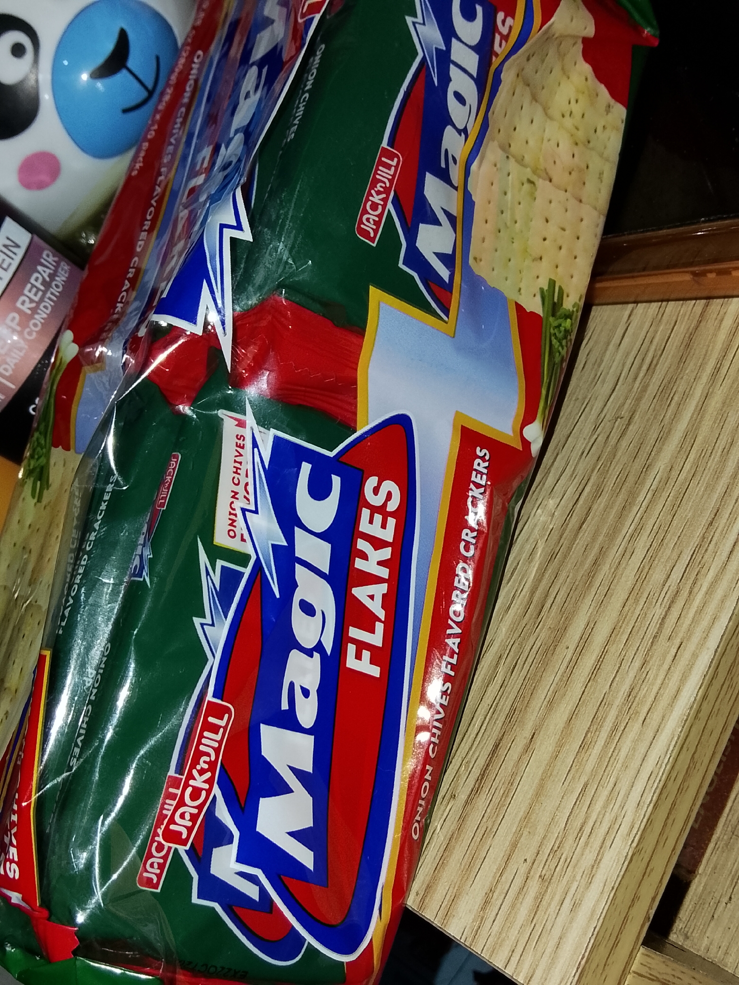 The image shows a close-up view of several packages of "Magic Flakes" crackers, specifically the Onion Chives flavor. The packages are predominantly green with red and blue accents, featuring prominent branding. Some of the cracker details, such as their perforated texture and small green chive pieces, are visible through the clear plastic packaging. The products are arranged in a way that suggests they are being displayed or stored, possibly in a retail setting or a home pantry. The background is a wooden surface with visible grain patterns. The lighting is somewhat dim, creating shadows that add depth to the scene. There are no people visible in the image. The environment appears to be indoors, with the wooden surface potentially being a tabletop or shelf. The context of Davao City, Philippines, is not discernible from the visual elements of the image itself.