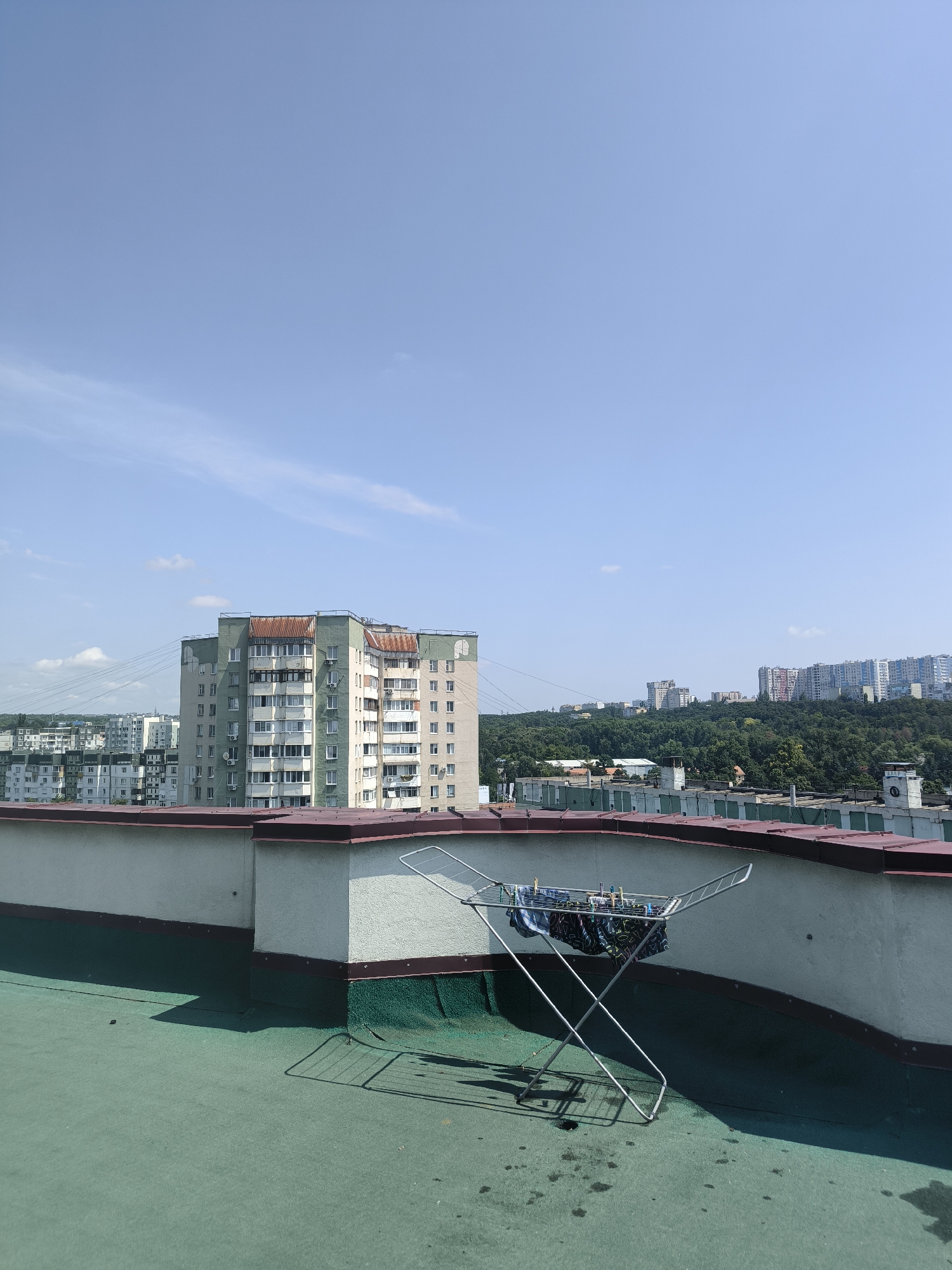 A high-angle shot captures a rooftop scene with a clear blue sky and a few wispy clouds. In the foreground, a foldable metal drying rack holds several pairs of colorful patterned underwear and some blue garments, secured with clothespins. The drying rack casts a distinct shadow on the green, textured rooftop, which shows some dark water stains and a small dark object, possibly a pebble.
The rooftop is bordered by a wall with a white textured finish and a dark red roof edge. Beyond this, a cityscape unfolds under the bright sunlight. A large, multi-story apartment building, painted in shades of green and light grey with a rusty-colored roof, dominates the middle ground. It features numerous windows and balconies. Further in the distance, more apartment buildings and other structures are visible, nestled against a backdrop of a lush, green, tree-covered hill. The overall impression is one of a sunny day in an urban environment, likely in the late morning or early afternoon given the shadows. There is no visible text in the image.