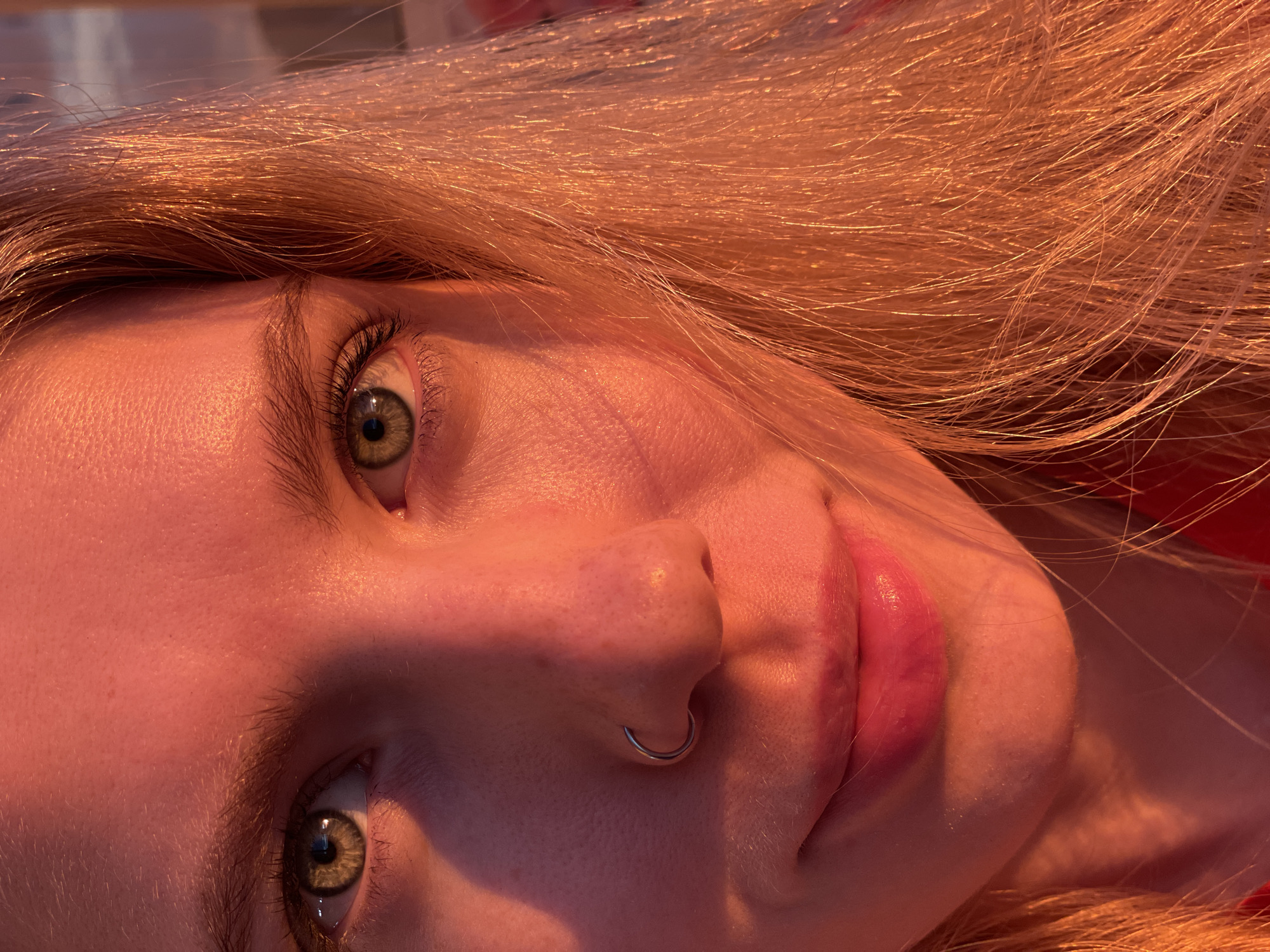 A close-up, eye-level, outdoor photograph features a young woman's face illuminated by warm, orange sunset light. Her face is turned slightly to the right, with her eyes looking directly at the camera. She has fair skin, light blonde hair, and striking green eyes with dark eyelashes. A silver septum ring is visible in her nose. Her lips are slightly parted and appear to be wearing pink lipstick. The background is out of focus and shows indistinct urban elements, suggesting an exterior city environment. The lighting indicates it is either sunrise or sunset. No specific landmarks in Izhevsk, Russia, are identifiable.