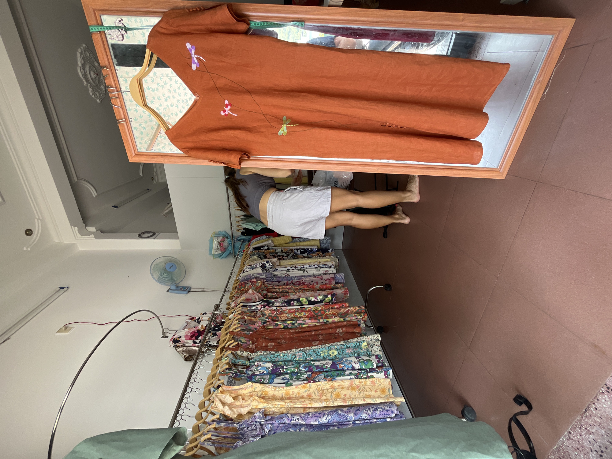 A rotated eye-level shot inside what appears to be a clothing shop in Bac Nha Trang Ward, Vietnam. A woman in a gray tank top and white shorts is bent over looking at stacked fabric rolls on a rack. An orange dress with dragonfly embroidery is hung in front of a mirror, the mirror is leaning against the rolls of fabric. The floor is composed of brown tiles. In the background, a fan is mounted on the white wall.