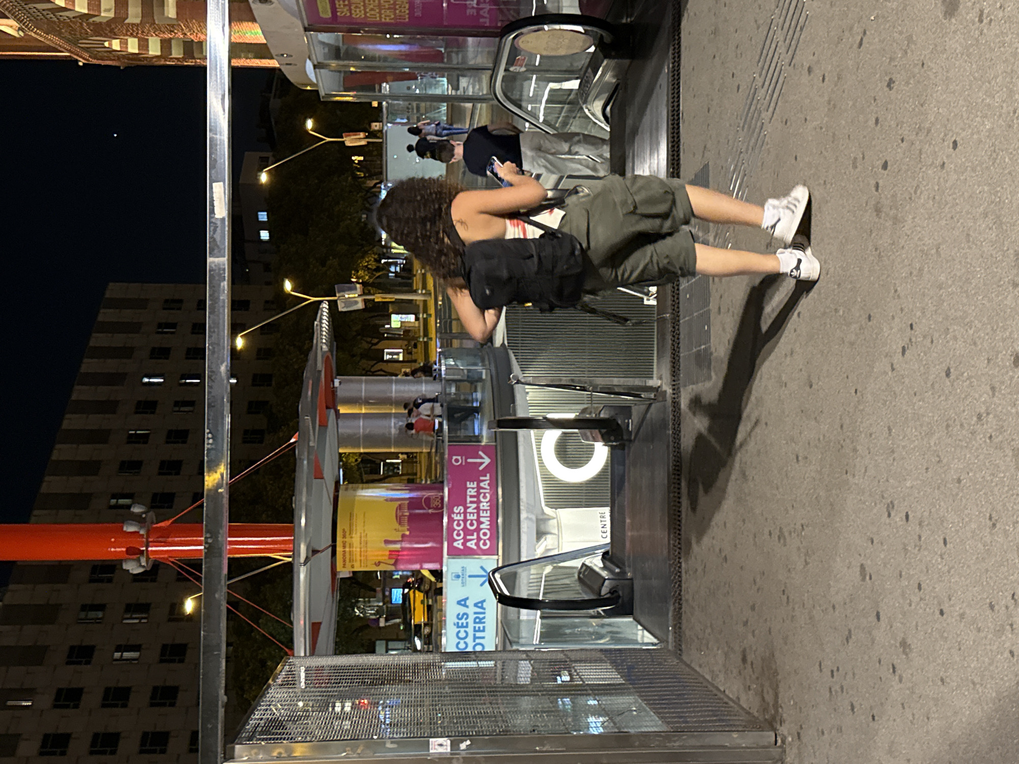 The image depicts a street scene at night in Barcelona, Spain, near an entrance to a commercial center. In the foreground, a young woman with curly dark hair and wearing a backpack, a patterned tank top, and olive green cargo shorts is walking away from the camera. She is holding a smartphone in her right hand. To her left, an escalator descends, marked with signs indicating "ACCÉS A LOTERIA" (Access to Lottery) and "ACCÉS AL CENTRE COMERCIAL" (Access to the Commercial Center). In the background, streetlights illuminate a building with multiple windows. Further back, other people are visible, some walking and others standing. The overall environment suggests an urban public space after dark.