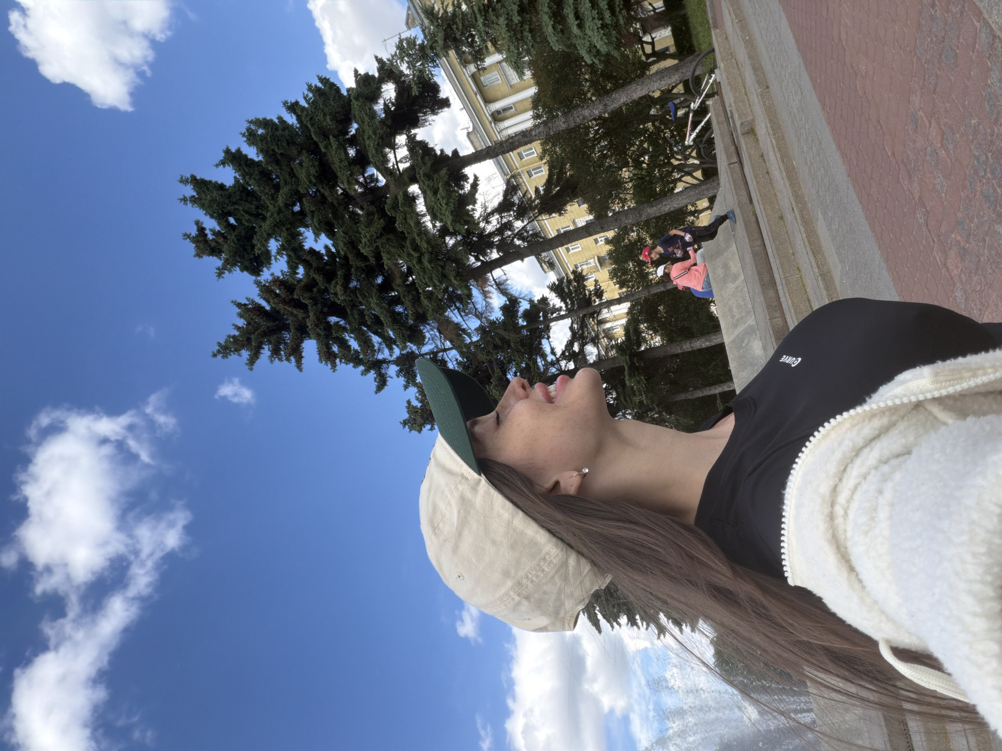 In an outdoor, daytime urban setting, likely Saint Petersburg, Russia, a young woman with long brown hair, wearing a beige baseball cap and a white zip-up jacket over a black t-shirt, is captured in a profile shot. Her eyes are closed, and she appears to be smiling, facing upwards towards a bright blue sky dotted with white clouds. Her left earlobe features a small hoop earring. In the background, tall evergreen trees stand prominently, with a yellow building featuring classical architecture and columns visible behind them. Several other individuals are present in the middle ground, including a person in a red cap and dark clothing running, and another individual in a pink jacket and white cap seated on a tiered stone structure. A bicycle is parked nearby. The ground is paved with red brick and grey concrete. A faint visual element suggesting a fountain or water spray is present to the far left of the frame.