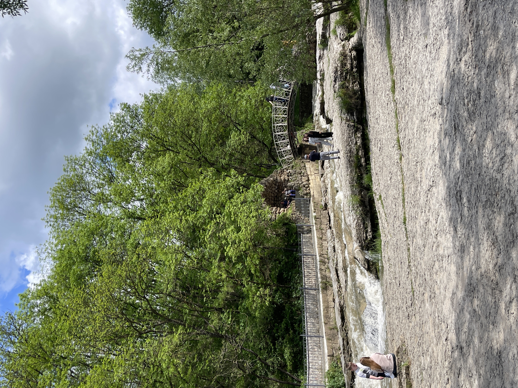 The image depicts a view in Kislovodsk, Russia, showcasing a natural stone terrace with a waterfall feature. Several people are present: some are positioned on an ornate metal bridge spanning the falls, while others are located on the stone terrace, observing or interacting with the environment. A smaller, enclosed structure is built into the stone near the bridge. Trees with lush green foliage dominate the left side of the frame, contrasting with the gray stone of the terrace. The sky is partially overcast.
