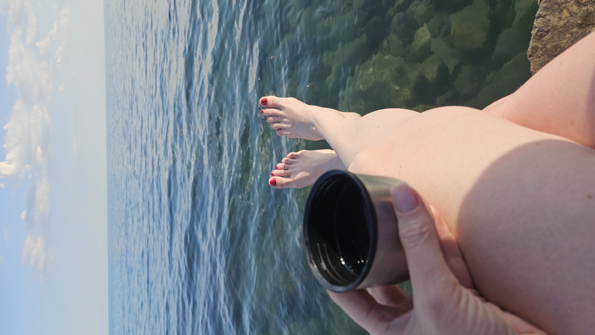 A first-person perspective shot shows a person's legs and feet dangling over the edge of what appears to be a stone pier. The person is holding a small, dark-colored insulated mug in their left hand. Their toenails are painted red. The legs and feet are illuminated by sunlight, creating shadows. The background is dominated by the calm, clear blue water of the sea, with visible stones on the seabed. In the distance, the horizon line separates the water from a pale blue sky with scattered white clouds. The location is identified as Dyurso, Russia.