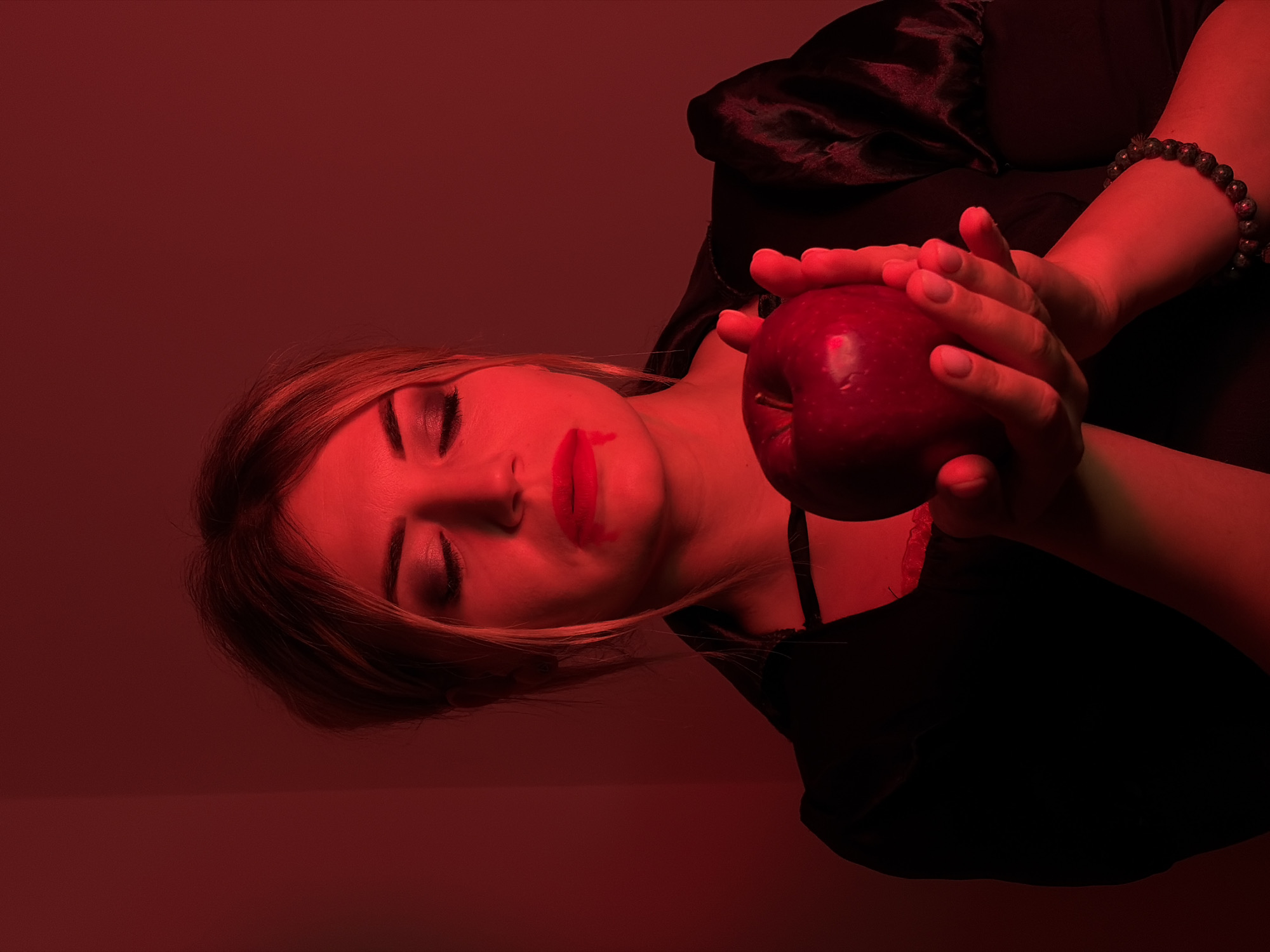 The image captures a person, likely a woman, in a dramatic, dark red-lit setting. She is depicted with her eyes closed, seemingly in a state of rest or contemplation. Her face is illuminated by the intense red light, highlighting red lipstick on her lips, which is visibly smeared down her chin. Her light-colored hair frames her face and spreads around her head.
She is wearing dark clothing, possibly a black top, and there's a dark, silky fabric draped around her shoulders and behind her head. Both of her hands are clasped around a vibrant red apple, holding it prominently in the center of the frame. A beaded bracelet is visible on her right wrist.
The entire scene is enveloped in a strong, monochromatic dark red light, casting deep shadows and creating an atmospheric and mysterious mood. The background is a plain wall, uniformly colored by the red lighting, indicating an indoor setting. There are no specific location cues visible, nor any discernible text within the image. The overall composition, along with the apple and the smeared lipstick, suggests themes often associated with fairy tales, temptation, or a moment of dramatic narrative.