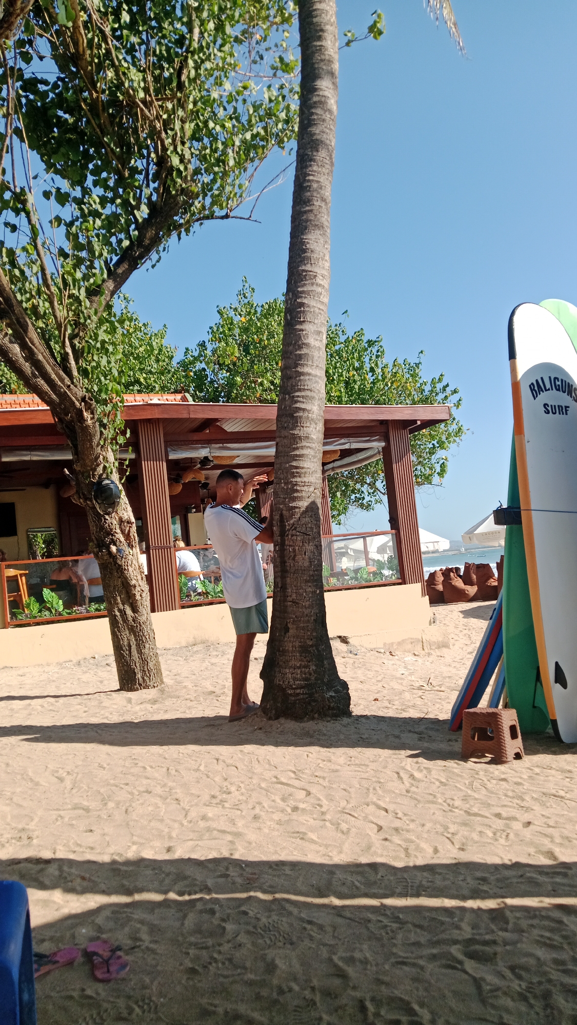 A man in a white t-shirt and shorts stands leaning against a palm tree on a sandy beach in Kuta, Indonesia. The man is looking towards the right and appears to be gesturing with his left hand. Behind him, a beachside building with a wooden roof and open sides is visible, with people seated inside. To the right of the man, several surfboards are propped up, one of which has "BALIGUNA SURF" written on it. The sky is clear and blue, and the sun casts shadows on the sand. In the foreground, a blue object and pink flip-flops are visible on the sand.