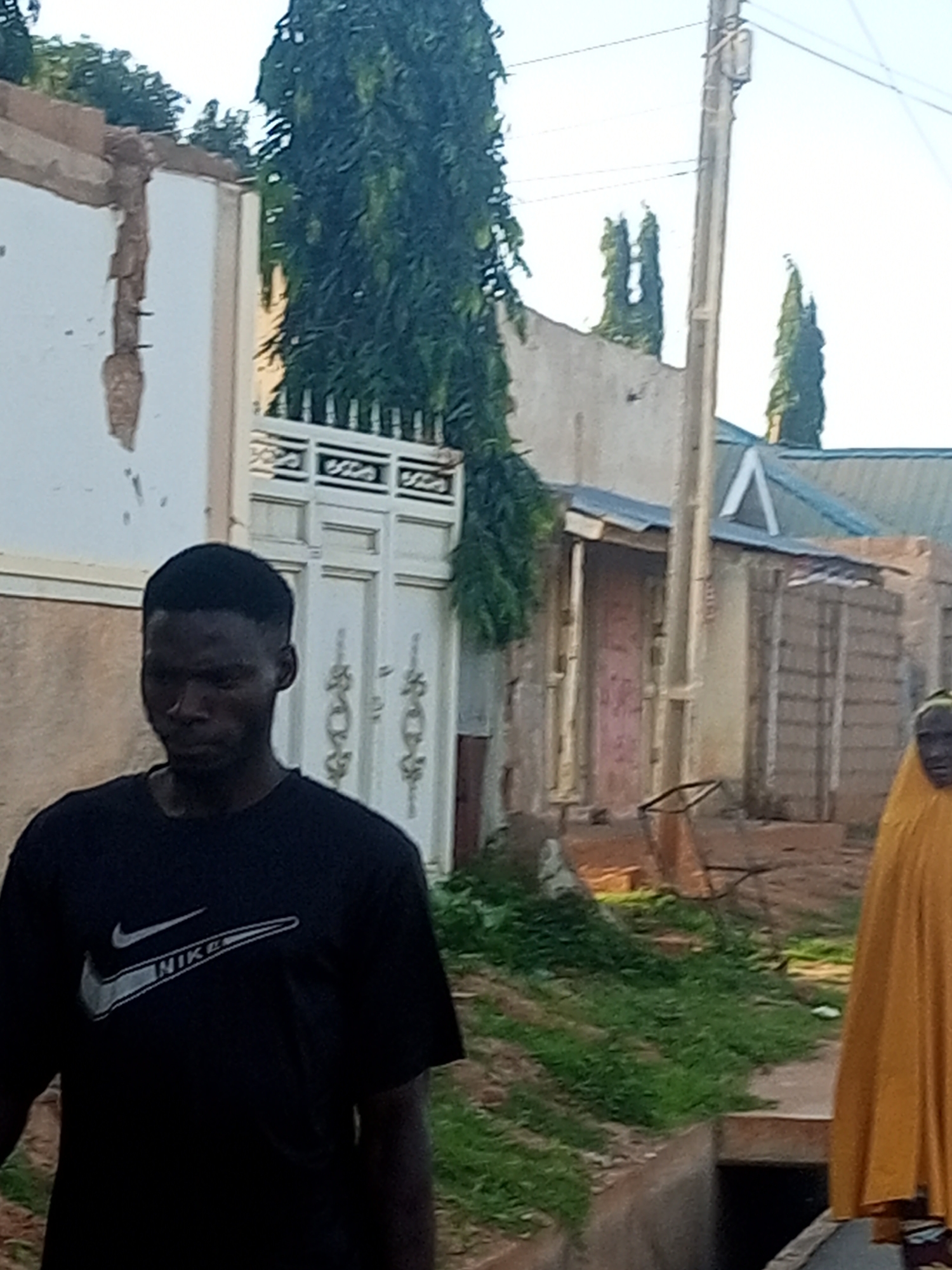 The image depicts an outdoor scene in Tempure, Nigeria. In the foreground, a young Black male is visible, wearing a black t-shirt with a white Nike logo. He is facing forward with his head slightly down, appearing to be in motion or pausing. To the right of the male, the lower part of a person wearing a bright yellow garment is visible, suggesting they are also in the scene, though their face and full body are not clearly shown.
The background features several structures, including buildings with textured walls and a white double door with decorative elements. A tall, slender tree with dense green foliage dominates the center-left of the frame, reaching towards the sky. A utility pole with wires is positioned slightly to the right of the tree. The environment appears to be a street or pathway with some areas of grass and debris, including what looks like an overturned metal object or frame. The overall lighting suggests it is daytime, with a clear sky. The buildings show signs of wear and tear, with some sections of plaster missing from the wall on the left.