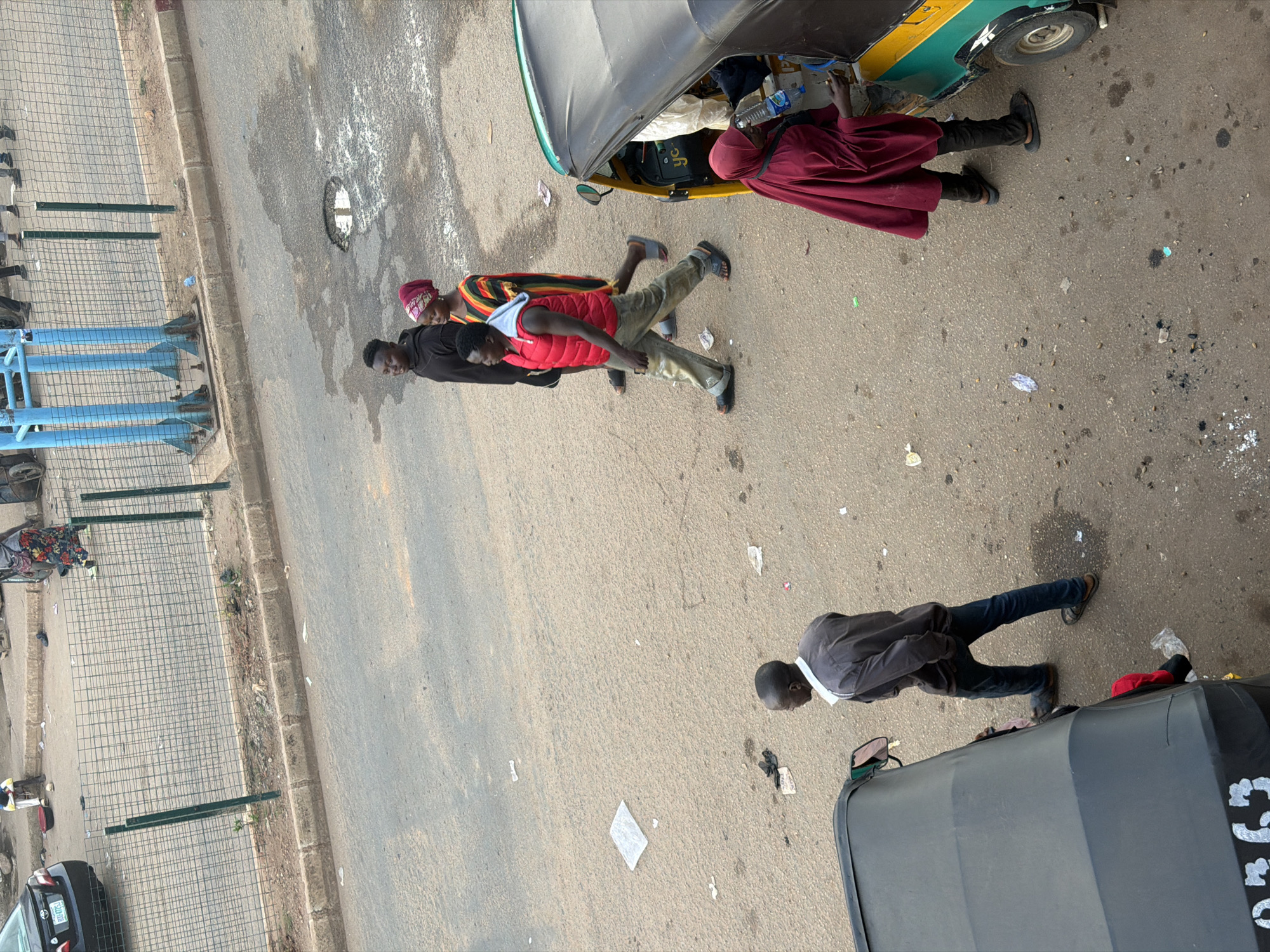 The image depicts an outdoor scene on a paved road, likely in Bukuru, Nigeria, based on the provided context. The perspective is from a high vantage point, looking down. Several people and vehicles are present.
In the foreground, a dark gray vehicle, possibly a minivan or an SUV, is partially visible from the top. To the left of this vehicle, a man wearing a dark gray long-sleeved shirt and blue jeans is walking away from the camera, with his head down. He is wearing dark shoes. To the right of him, partially obscured by the vehicle, is the rear of another dark vehicle, possibly a utility vehicle or a modified car, with the letters "C 135" visible in white spray paint on its side.
Further up the road, a bright yellow and green three-wheeled vehicle, commonly known as a "keke napep" or "tricycle taxi," is parked. A person wearing a maroon head covering and a long maroon garment is standing by the open door of the keke, appearing to interact with someone inside or nearby. Another person, wearing a dark top and a red vest, is also near the keke, with their back to the camera. Behind them, another person is visible wearing a colorful striped top and a red head covering.
To the far left of the image, a wire mesh fence structure is set up along the roadside. Beyond the fence, a sidewalk and some greenery are visible. Scattered on the road are various pieces of litter, suggesting a public area. The lighting suggests it is daytime, with bright sunlight casting shadows, but the overall weather appears to be clear. The overall atmosphere seems to be that of a busy street scene with economic activity.