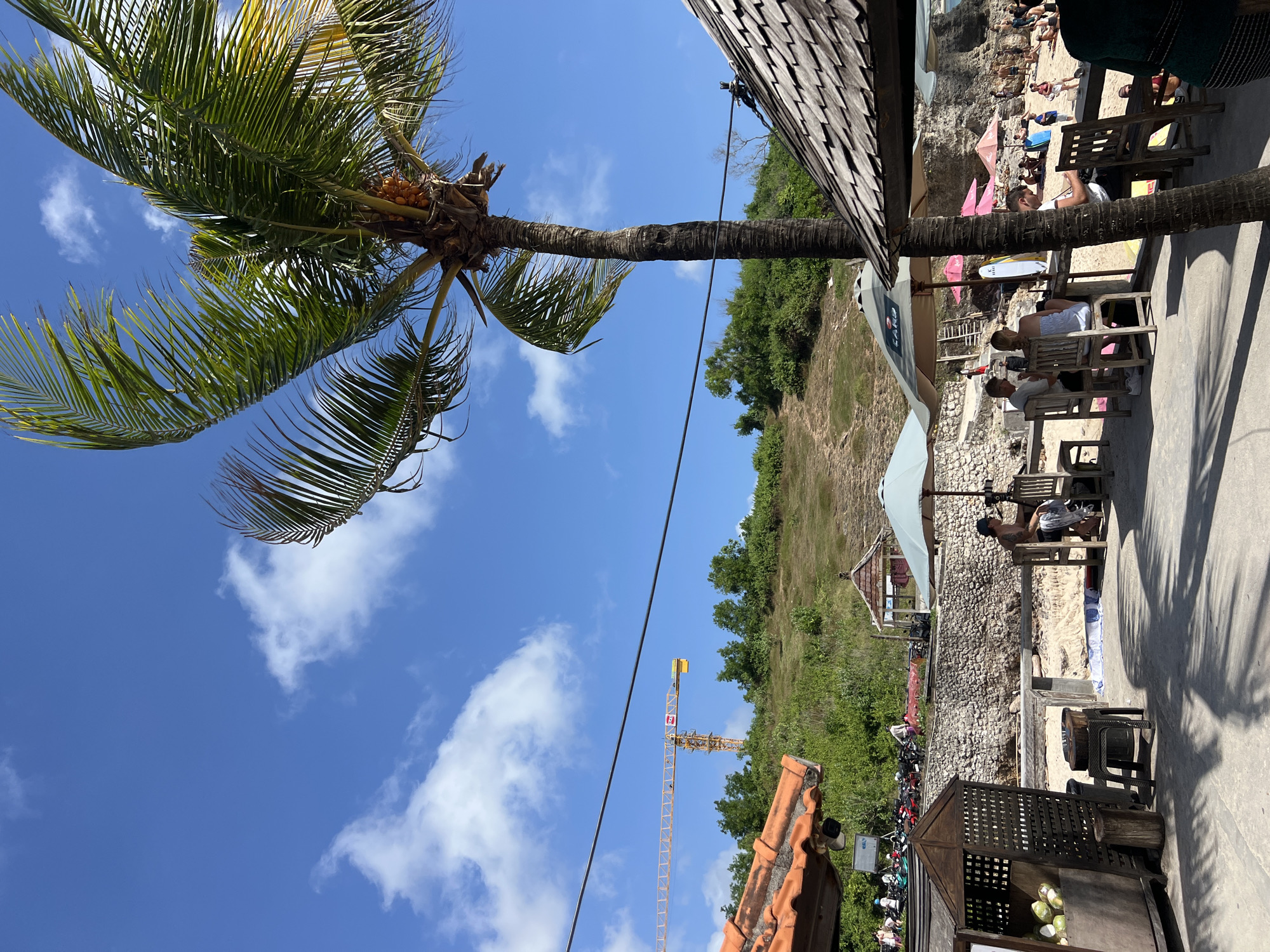 The scene depicts a beachside location in Kuta Selatan, Indonesia. A palm tree with coconuts is prominent in the foreground against a clear blue sky with scattered clouds. To the right, there's a partially visible wooden structure, possibly a building or roof. Below the building and receding into the distance, the scene transitions to a coastal area with a beach. People are visible sunbathing and sitting at tables under umbrellas. Further back, there's a hillside with green vegetation and a construction crane. Various motorbikes and a small stall are noticeable. The general atmosphere is that of a relaxed beach environment.